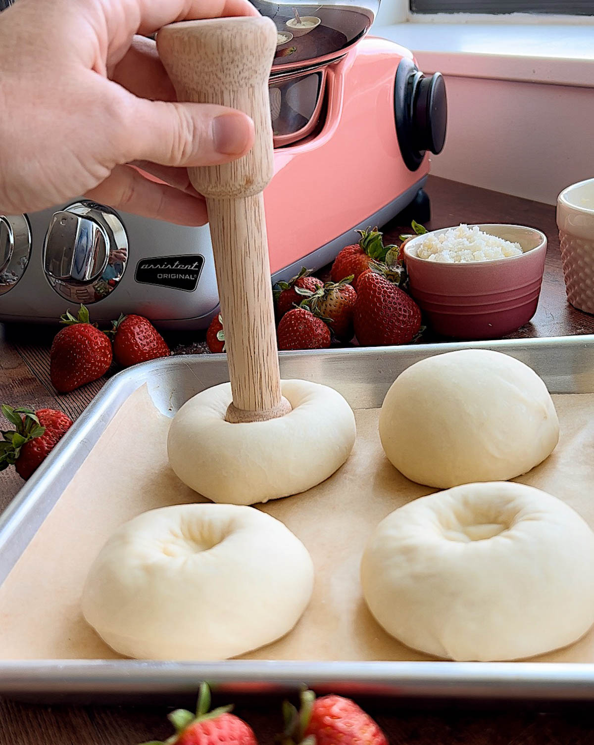 A hand holding a wooden tamper, pressing a indentation into the center of a roll, on a baking sheet lined with parchment paper.