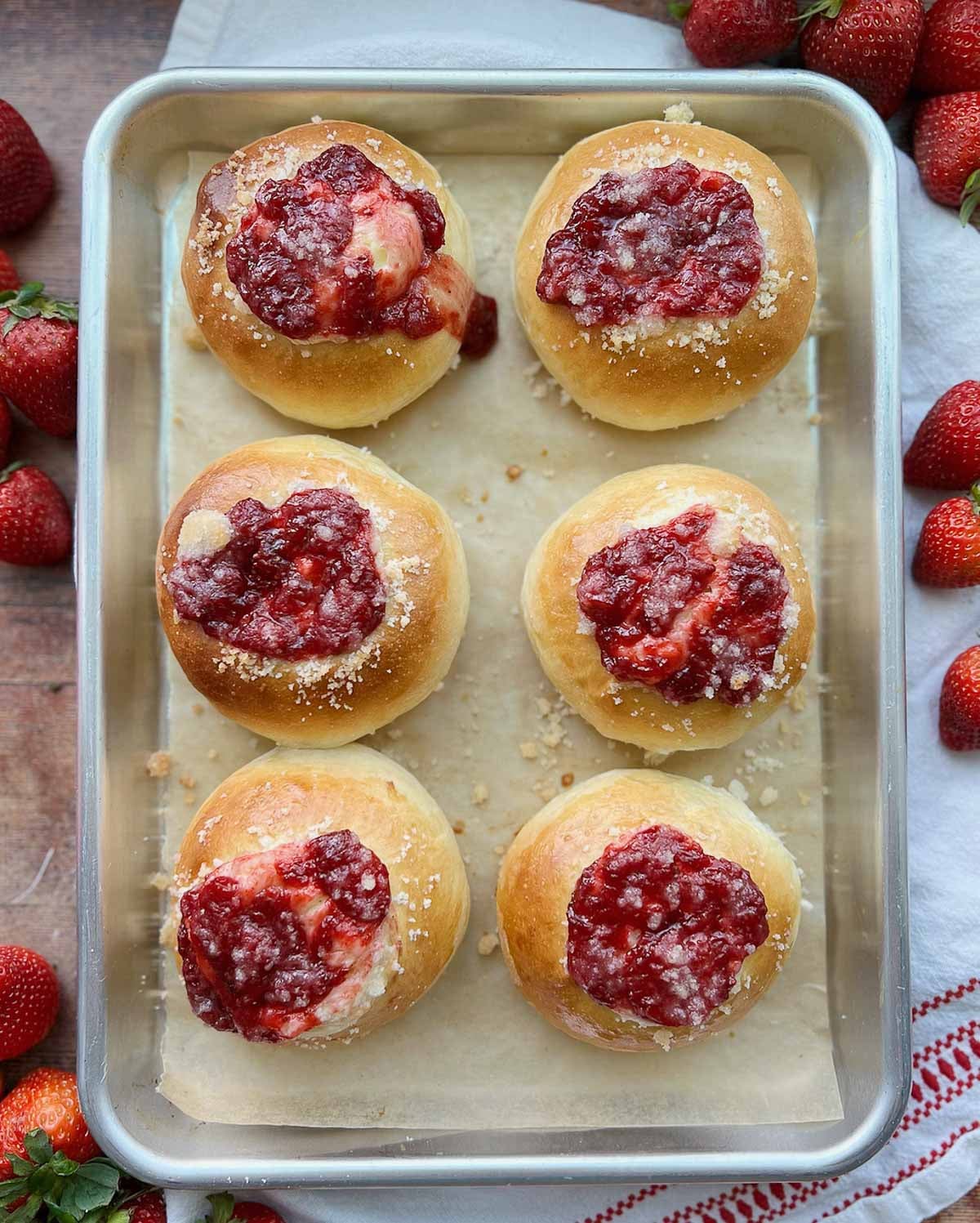 Baked strawberry cream cheese rolls on a baking with lined with parchment, and strawberries on the counter around it.
