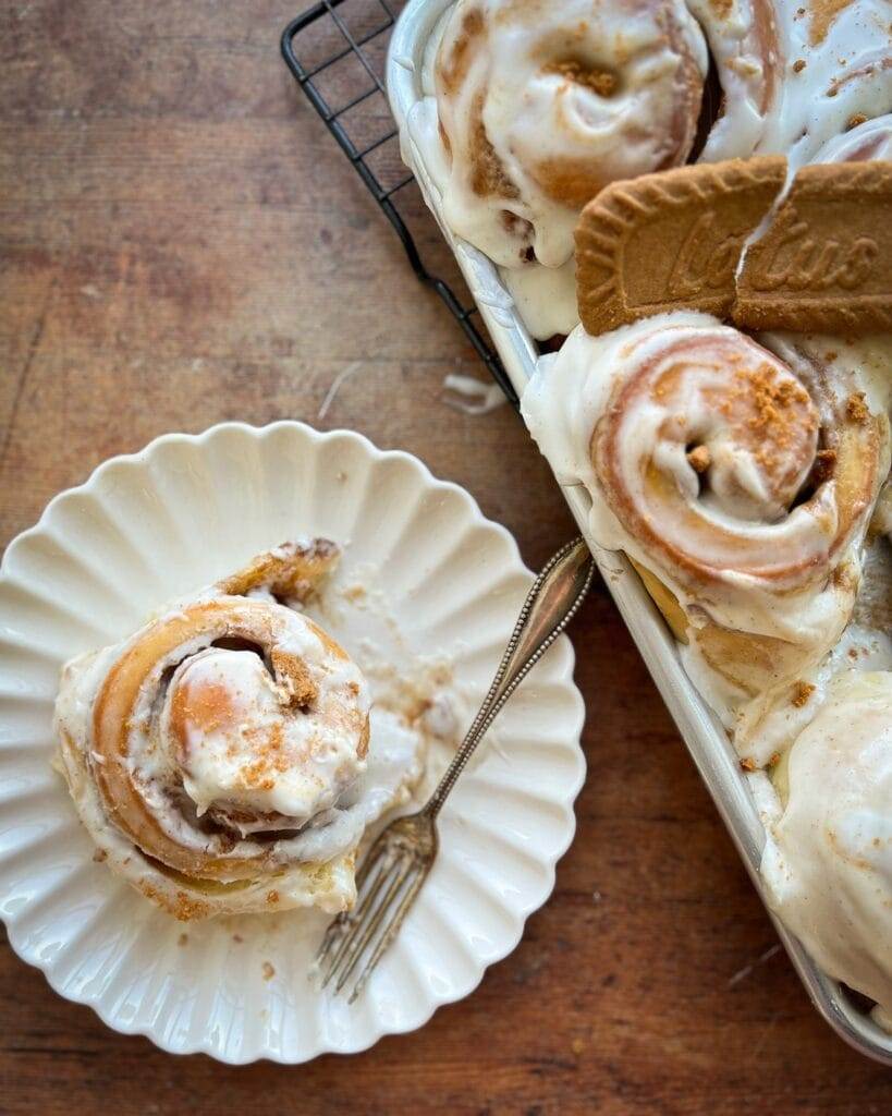 A biscoff cinnamon roll on a plate next to a pan of cinnamon rolls.