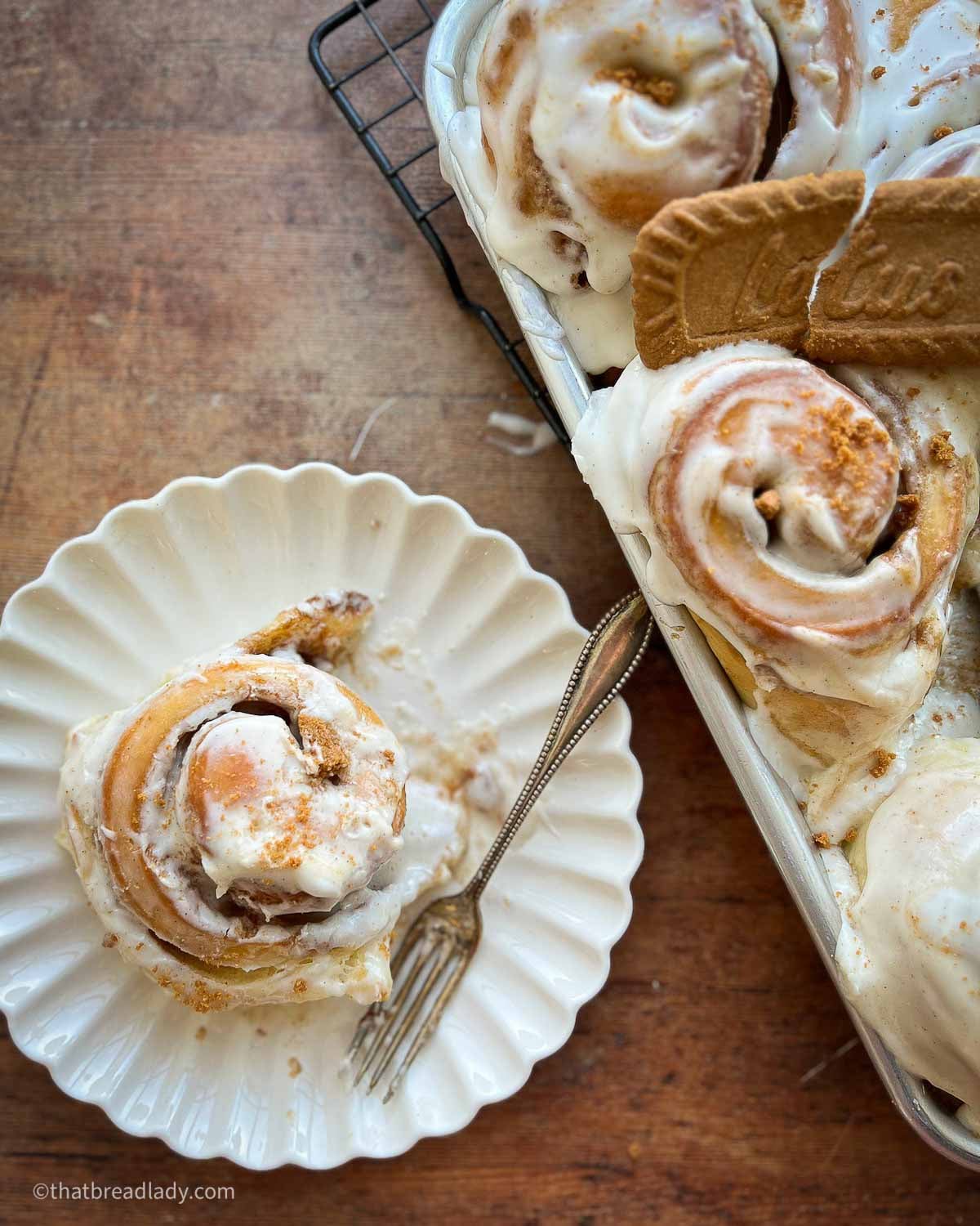 A Biscoff cinnamon roll on a dessert plate with a fork, and a pan of more rolls.