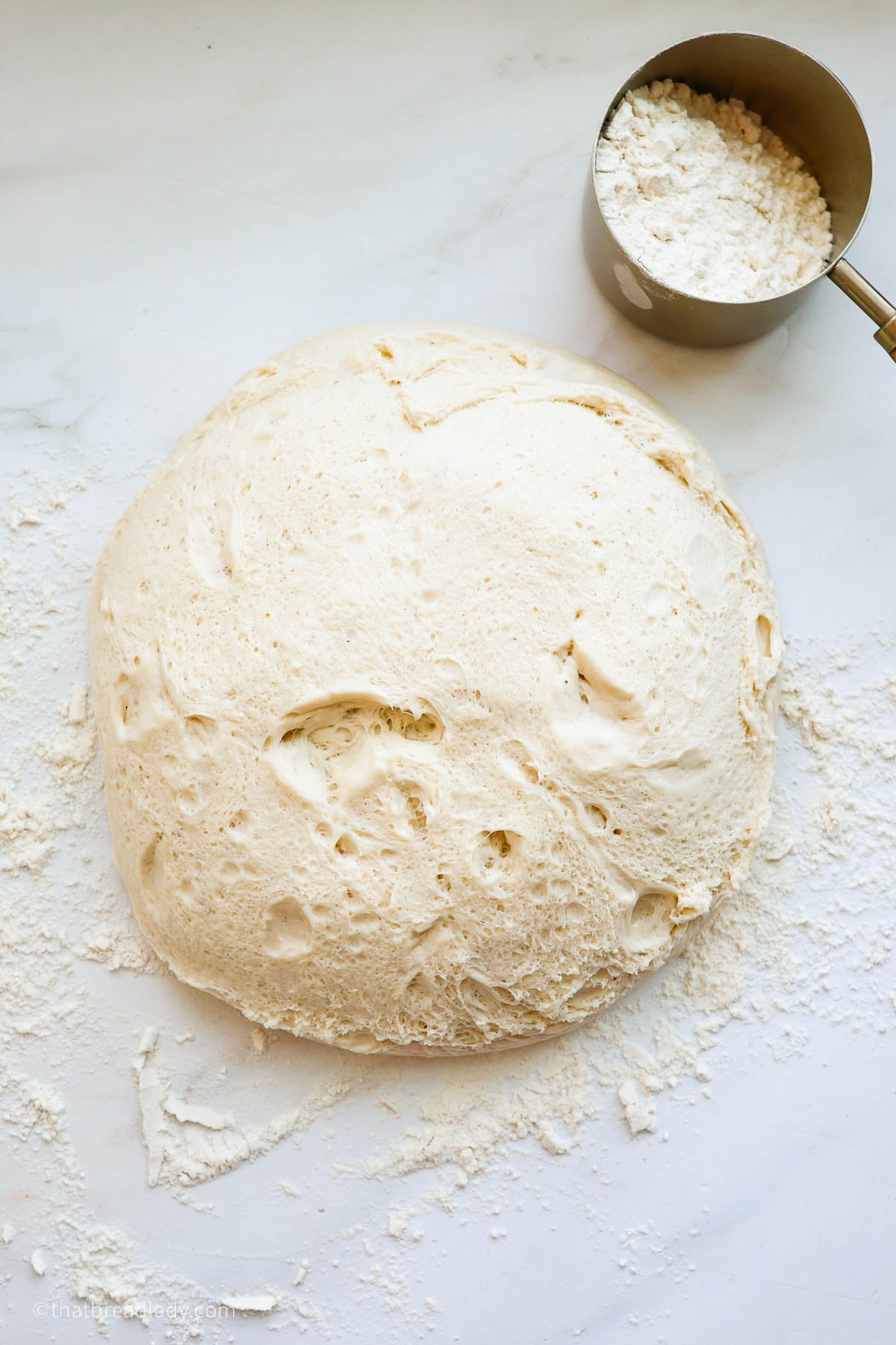 A ball of risen dough, on a floured countertop, with a measuring cup full of flour.