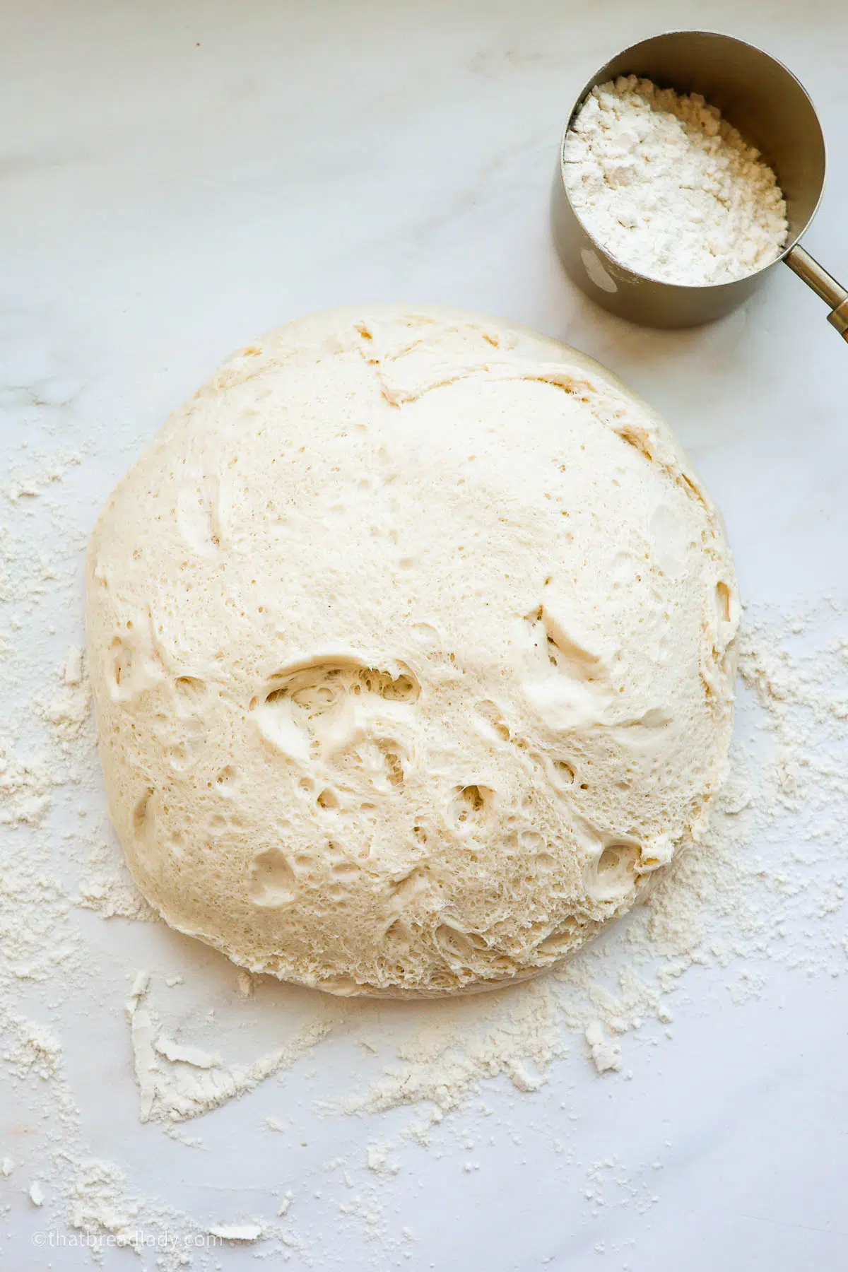 A ball of risen dough, on a floured countertop, with a measuring cup full of flour.