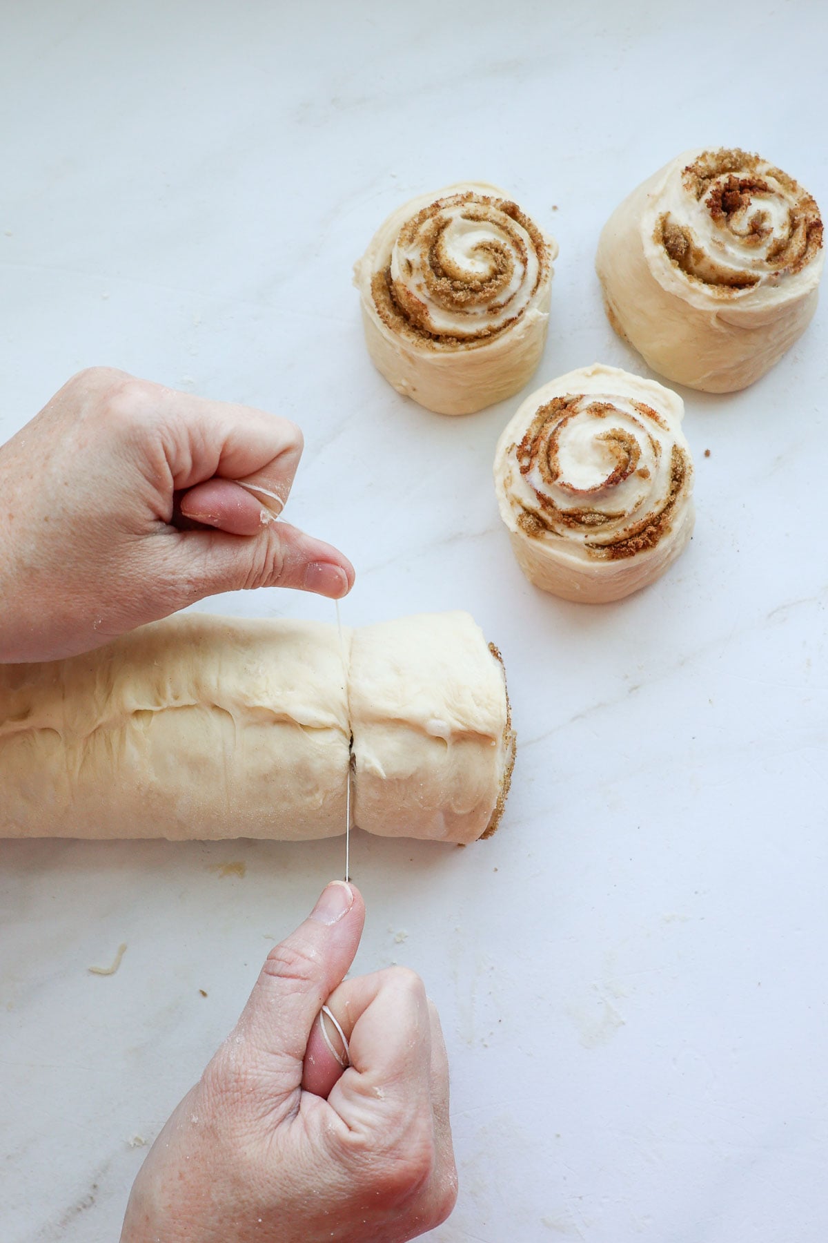 Two hands, using dental floss to cut a Biscoff cinnamon roll from a log of dough, with three already cut Biscoff cinnamon rolls on the work surface.