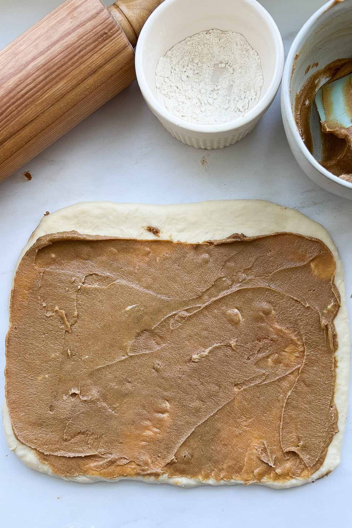A piece of cinnamon roll dough, rolled out to a rectangle, with Biscoff filling spread over it, with a clean boarder at the top, a rolling pin, and a bowl of flour.