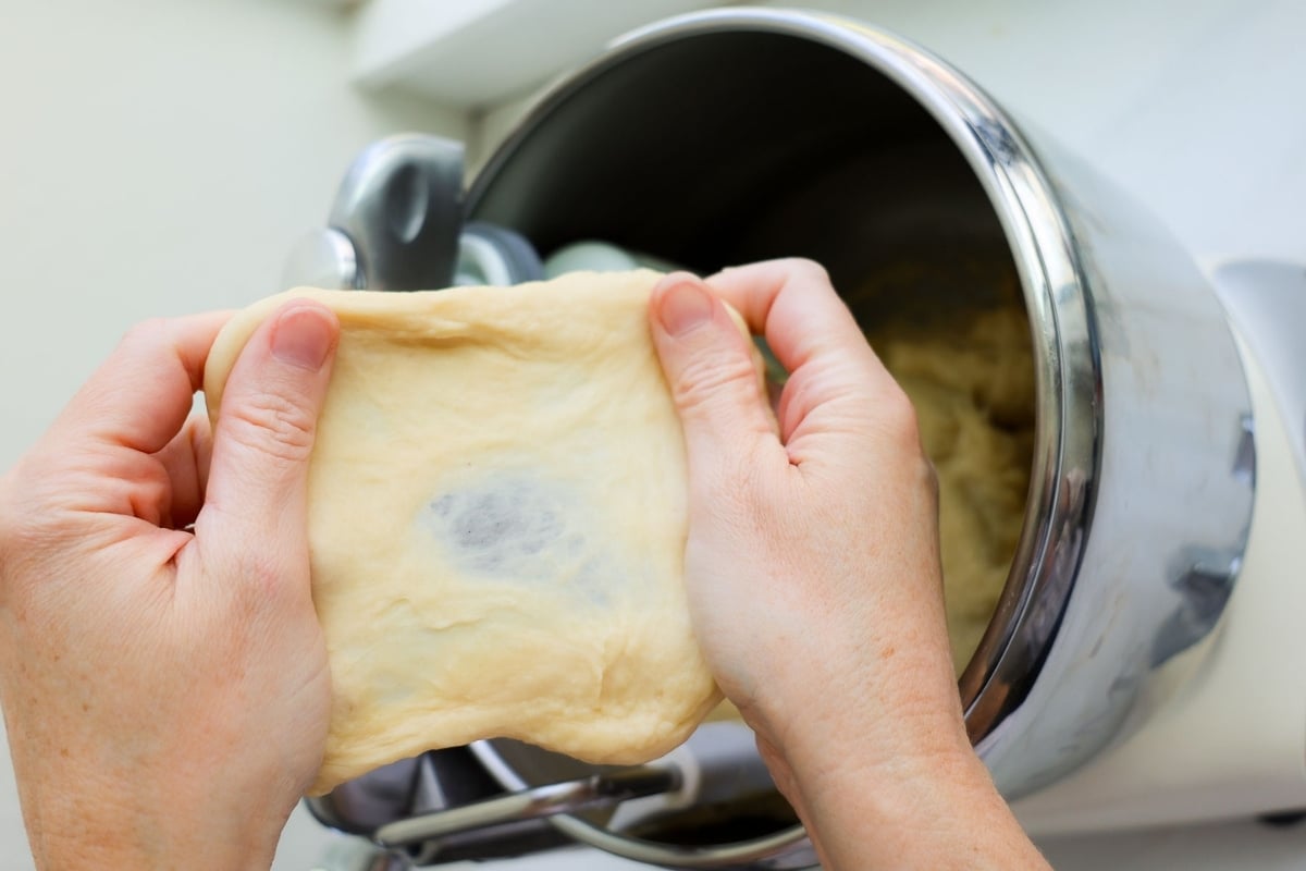 Two hands holding a piece of Biscoff cinnamon roll dough and stretching it apart, with the center of the dough translucent.