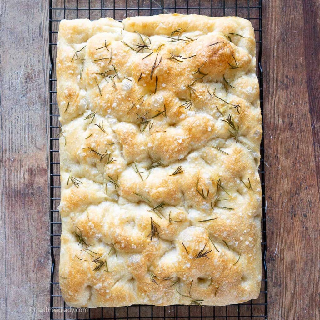 A baked focaccia, topped with flaky salt and rosemary, resting on a wire cooling rack.