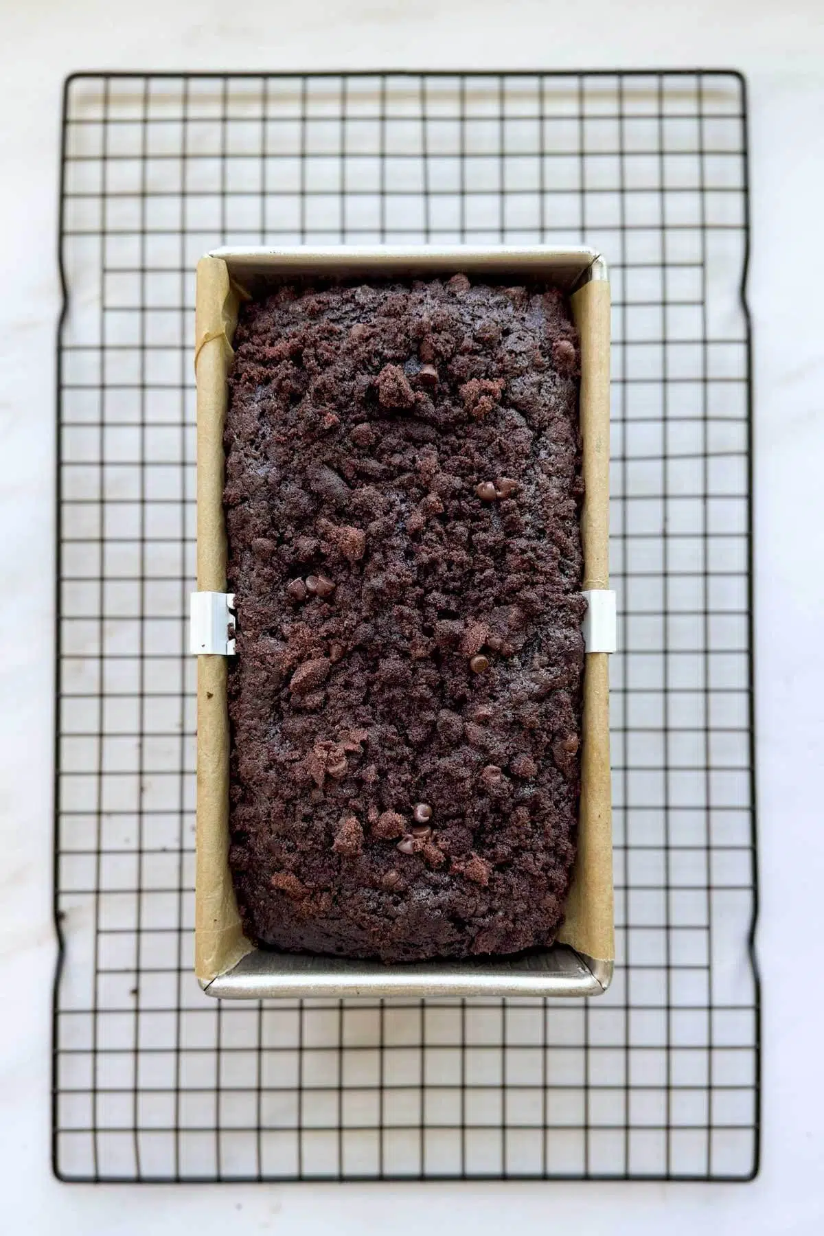 A baked loaf of double chocolate zucchini bread in a loaf pan lined with parchment paper, resing on a wire cooling rack.