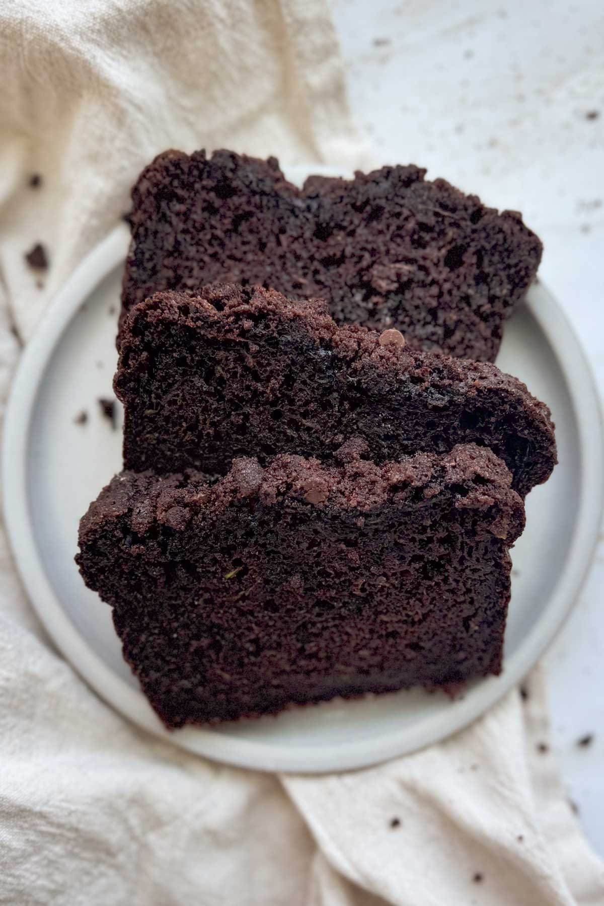 Three slices of double chocolate zucchini bread on a white plate with a white linen napkin underneath it.