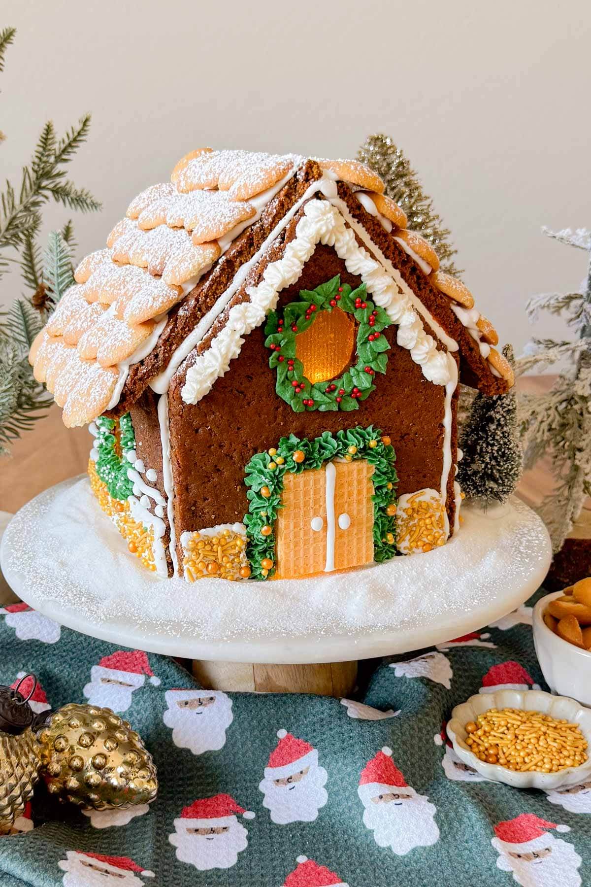 A gingerbread house decorated with Nilla Wafers and powdered sugar on the roof, a candied window with lights glowing inside, and frosting piped on the front as garland and a wreath.