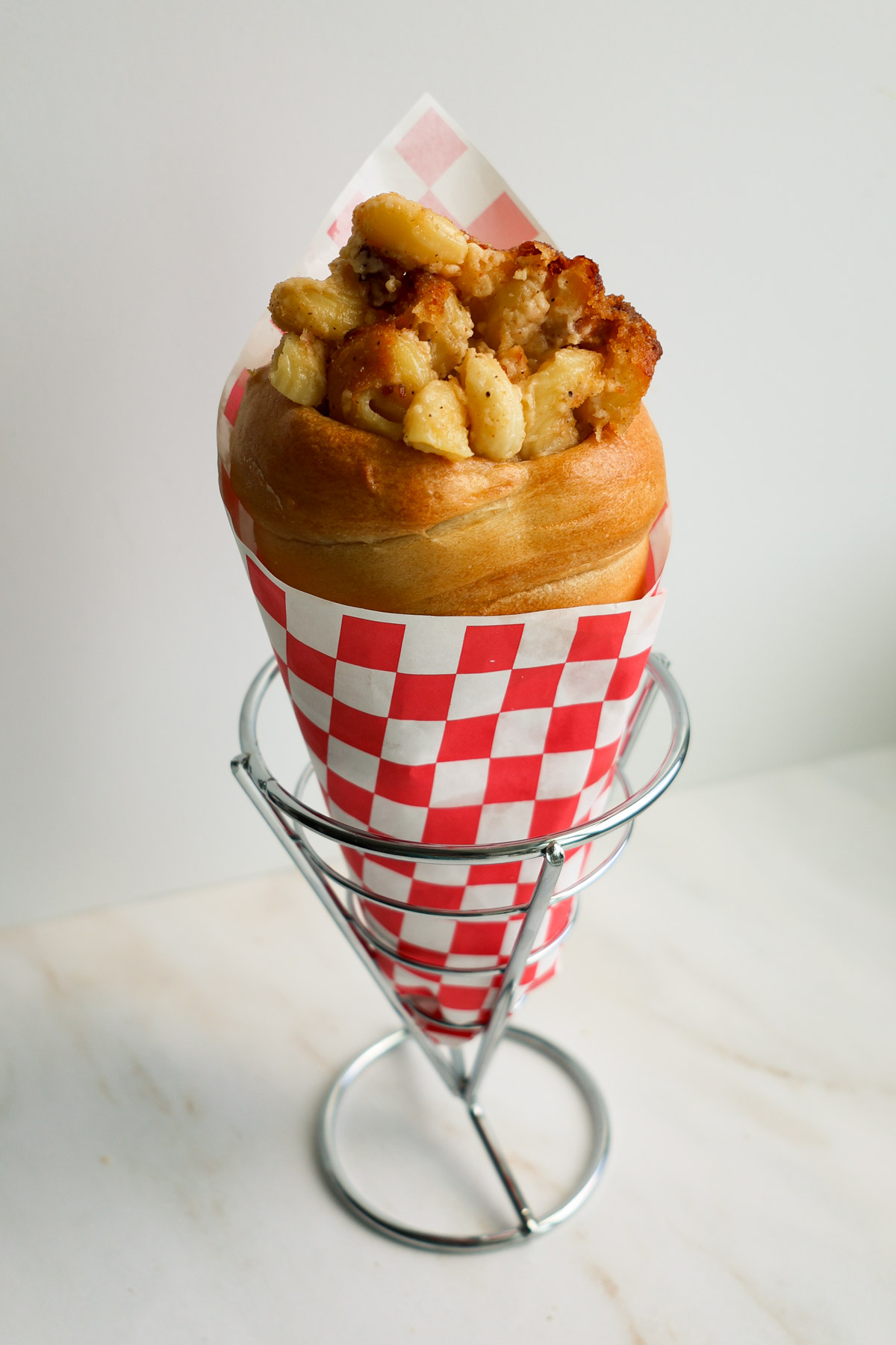 A bread cone with bacon mac and cheese, wrapped in a red and white checkered paper, resting in a cone basket.