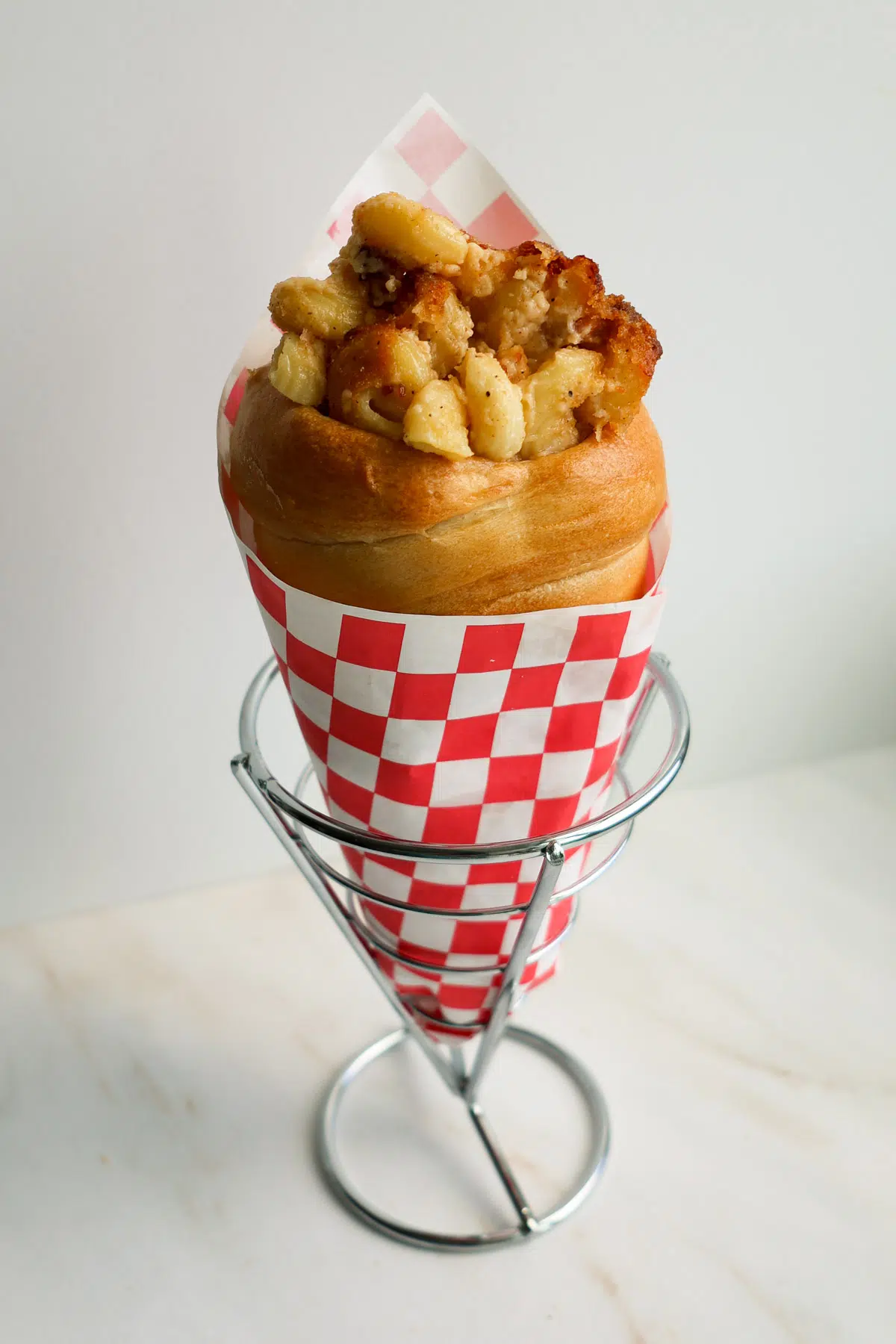 A bread cone with bacon mac and cheese, wrapped in a red and white checkered paper, resting in a cone basket.