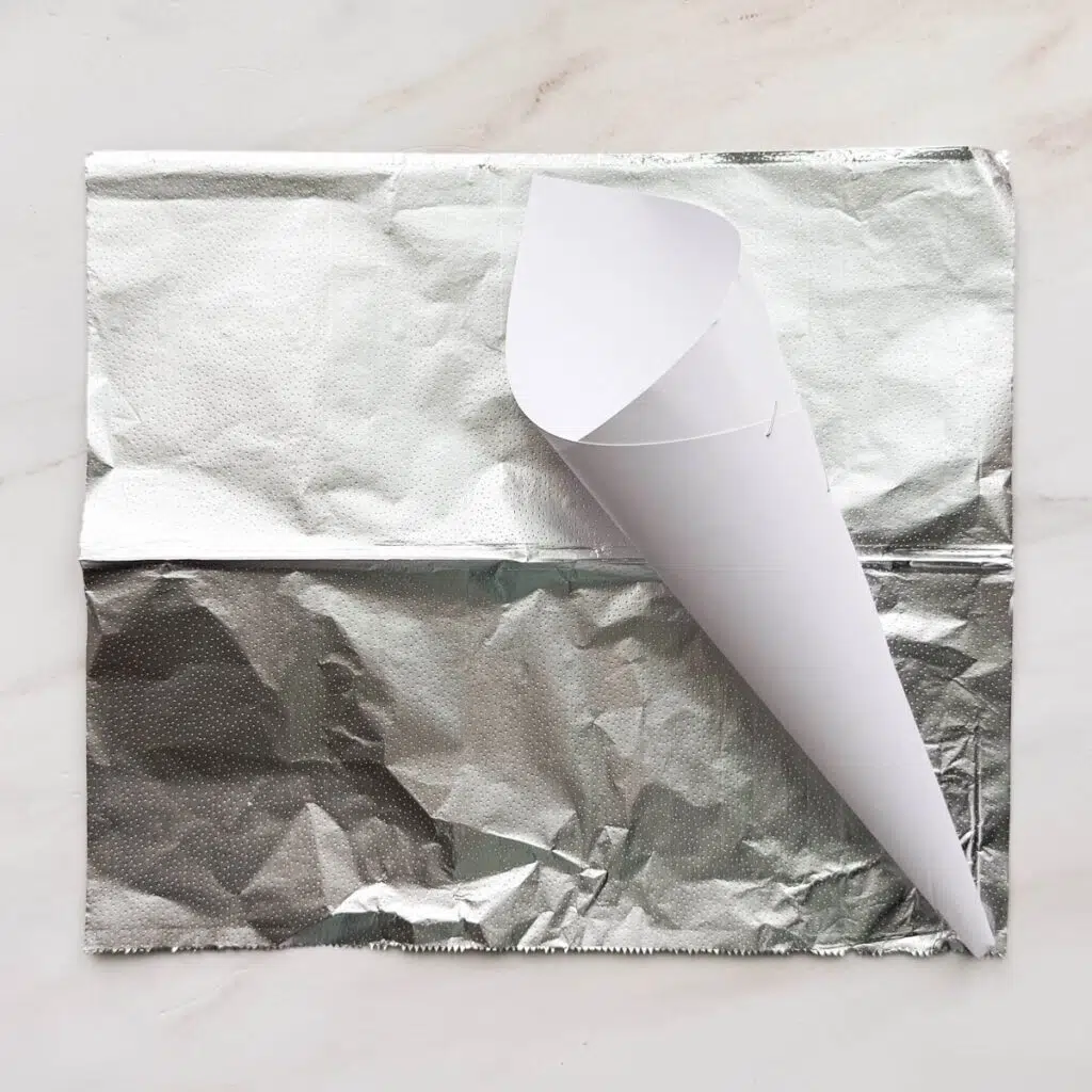 A white piece of paper rolled up in a cone shape, laying on top of a sheet of aluminum foil.