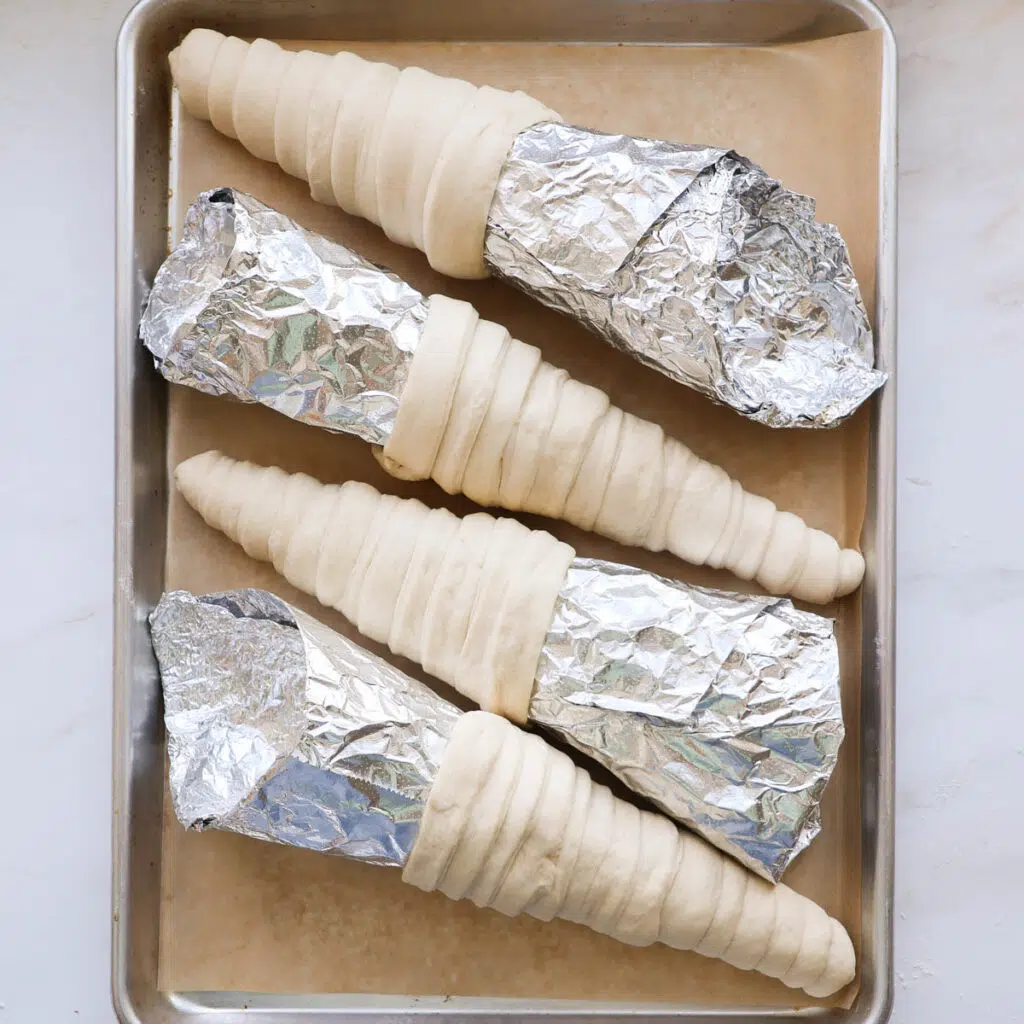 Four aluminum foil cone molds with bread dough wrapped around them, on a baking sheet lined with parchment paper.