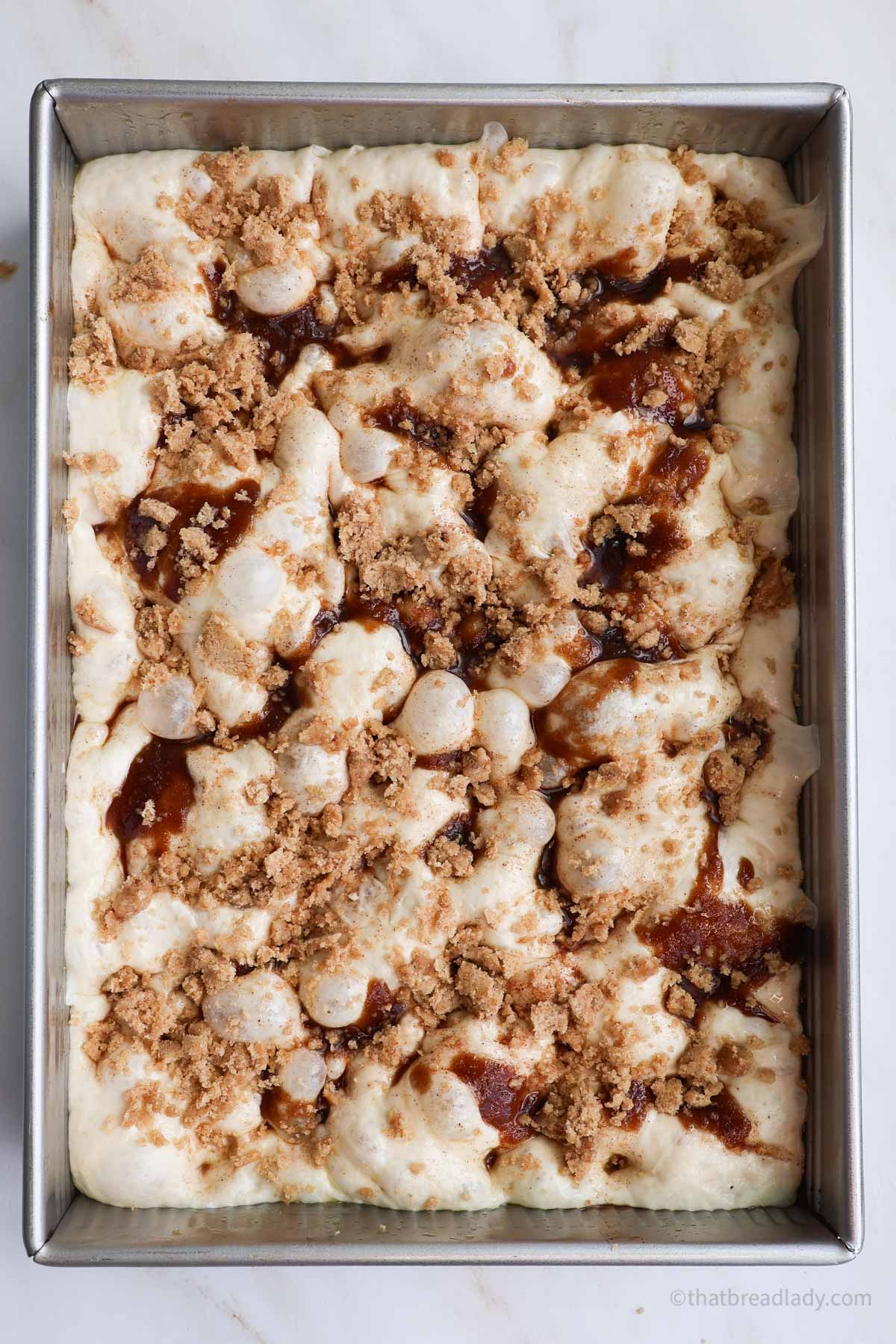 A pan of Cinnamon Roll Focaccia dough, dimpled, topped with cinnamon butter filling and a crumb topping.