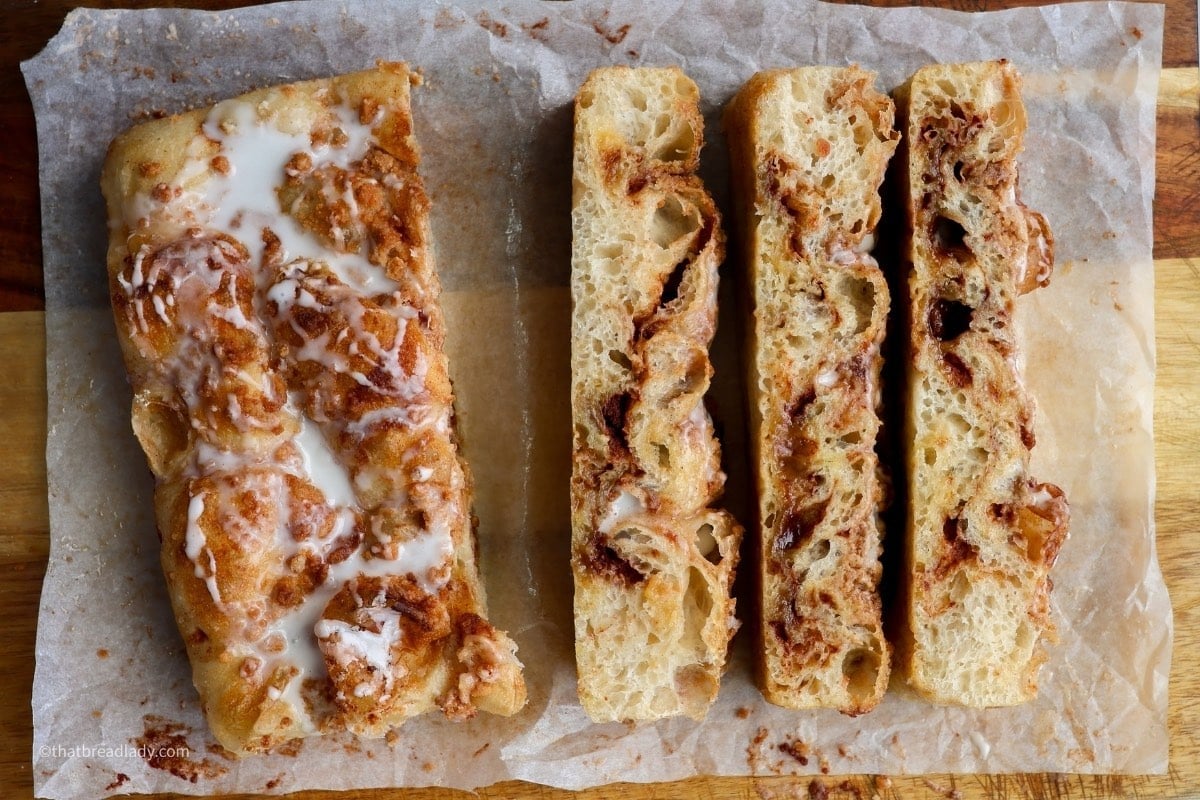Sliced cinnamon roll focaccia, with a vanilla icing, resting on a wood cutting board that is lined with parchment paper.