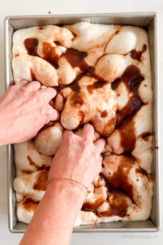 Two hands dimpling the dough of cinnamon roll focaccia, with cinnamon butter filling on top of the dough.
