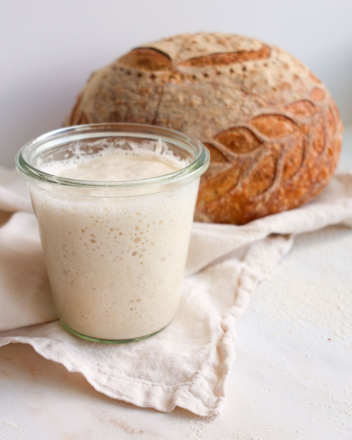 A jar of sourdough starter with a loaf of bread in the background.