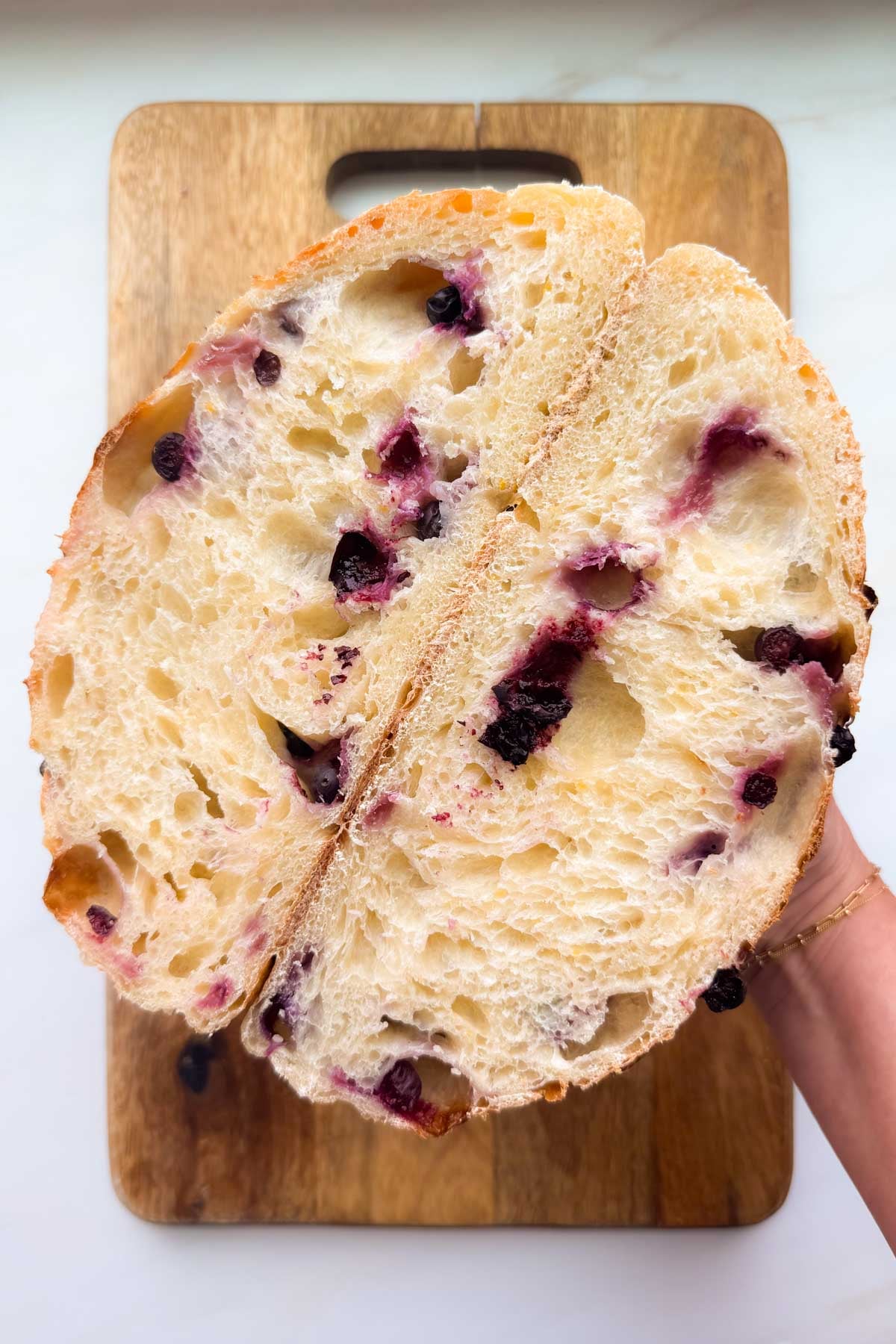 Cross-section of a perfectly baked lemon blueberry sourdough bread, held in a hand above a wooden cutting board.