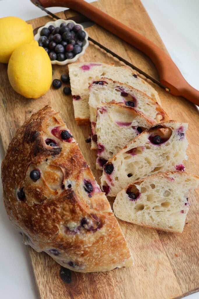 A baked blueberry lemon sourdough bread cut in half with examples of slices of the bread on a wood cutting board with a serrated bread knife, fresh lemons, and a cup of blueberries.