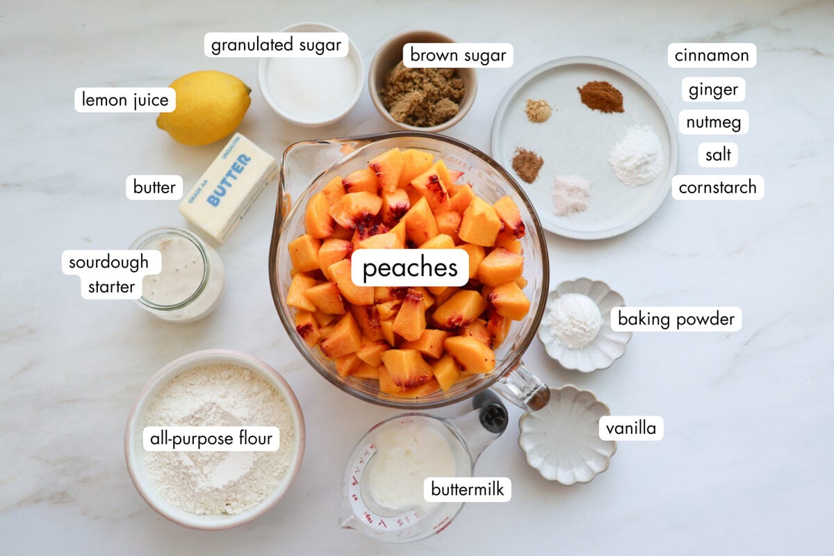 All the ingredients for sourdough peach cobbler, measured out, including chopped peaches, brown sugar, granulated sugar, a lemon, butter, sourdough starter, flour, buttermilk, vanilla, baking powder, cinnamon, ginger, nutmeg, and cornstarch.