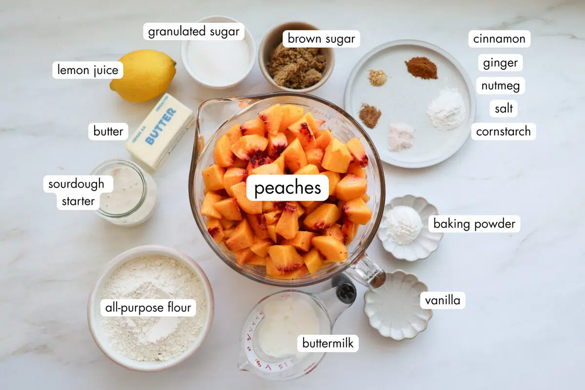 All the ingredients for sourdough peach cobbler, measured out, including chopped peaches, brown sugar, granulated sugar, a lemon, butter, sourdough starter, flour, buttermilk, vanilla, baking powder, cinnamon, ginger, nutmeg, and cornstarch.