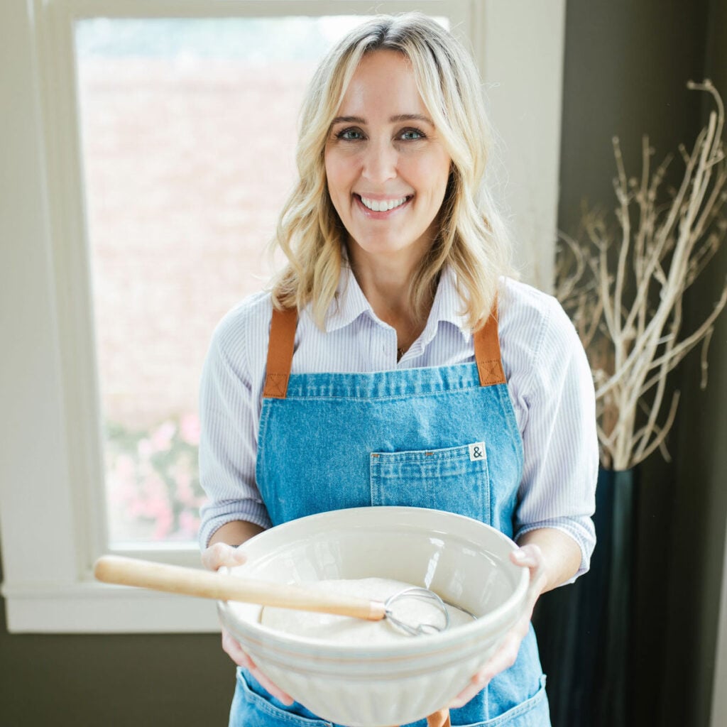 Heather from That Bread Lady, wearing an apron, holding a large bowl with bread dough in it and a Danish whisk resting on top.