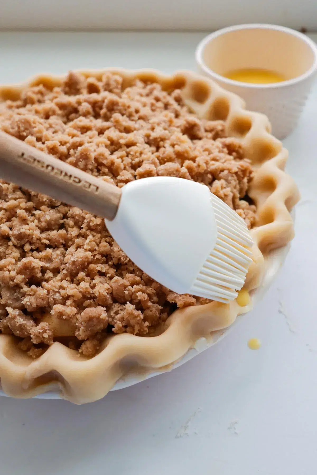 A pastry brush brushing the crust of an unbaked apple crumble pie with egg wash.