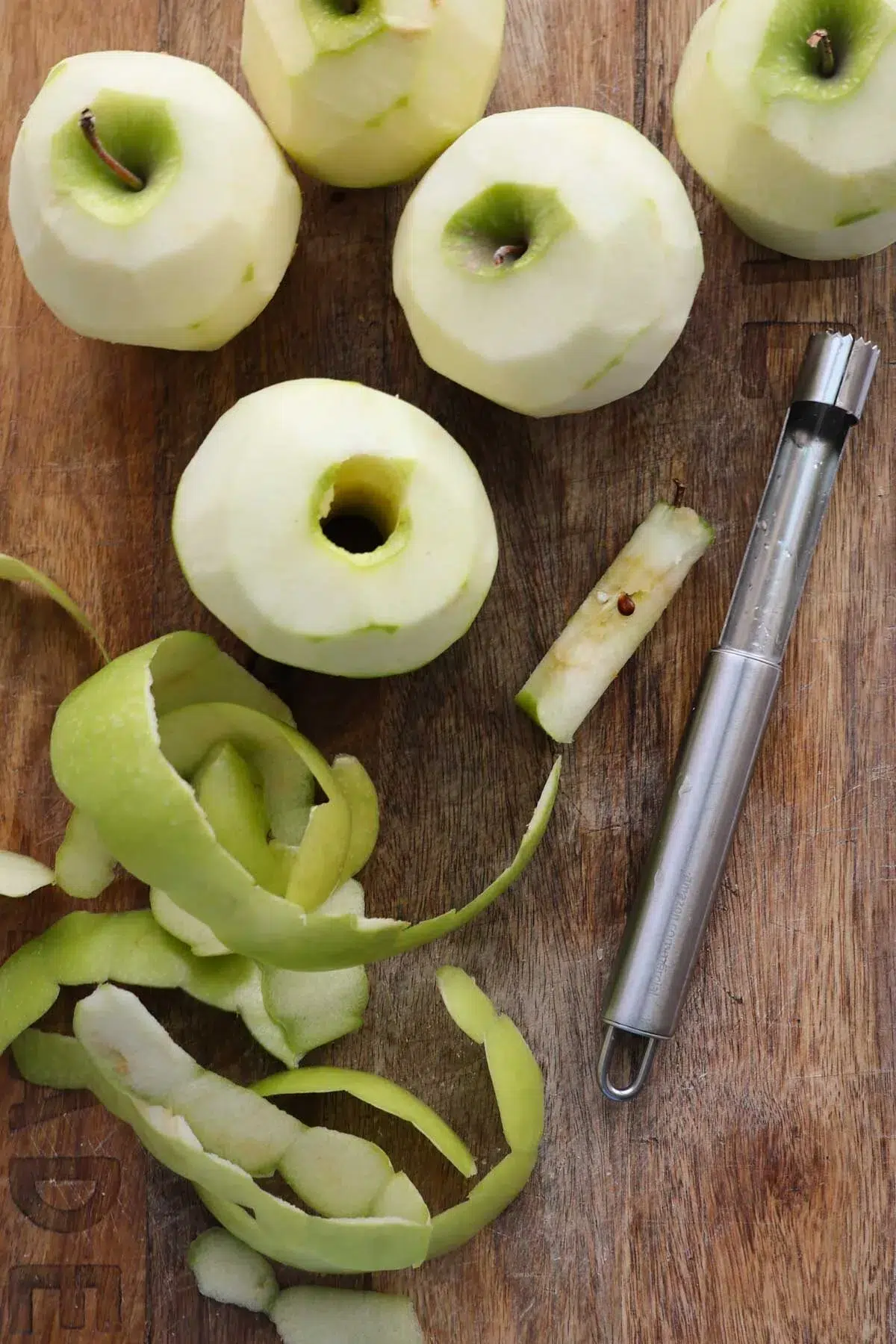 Several Granny Smith apples, cored and peeled, with peels and a corer on the wood cutting board they are on.