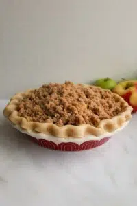 An unbaked apple crumble pie, topped with crumb topping, and two apples next to it.