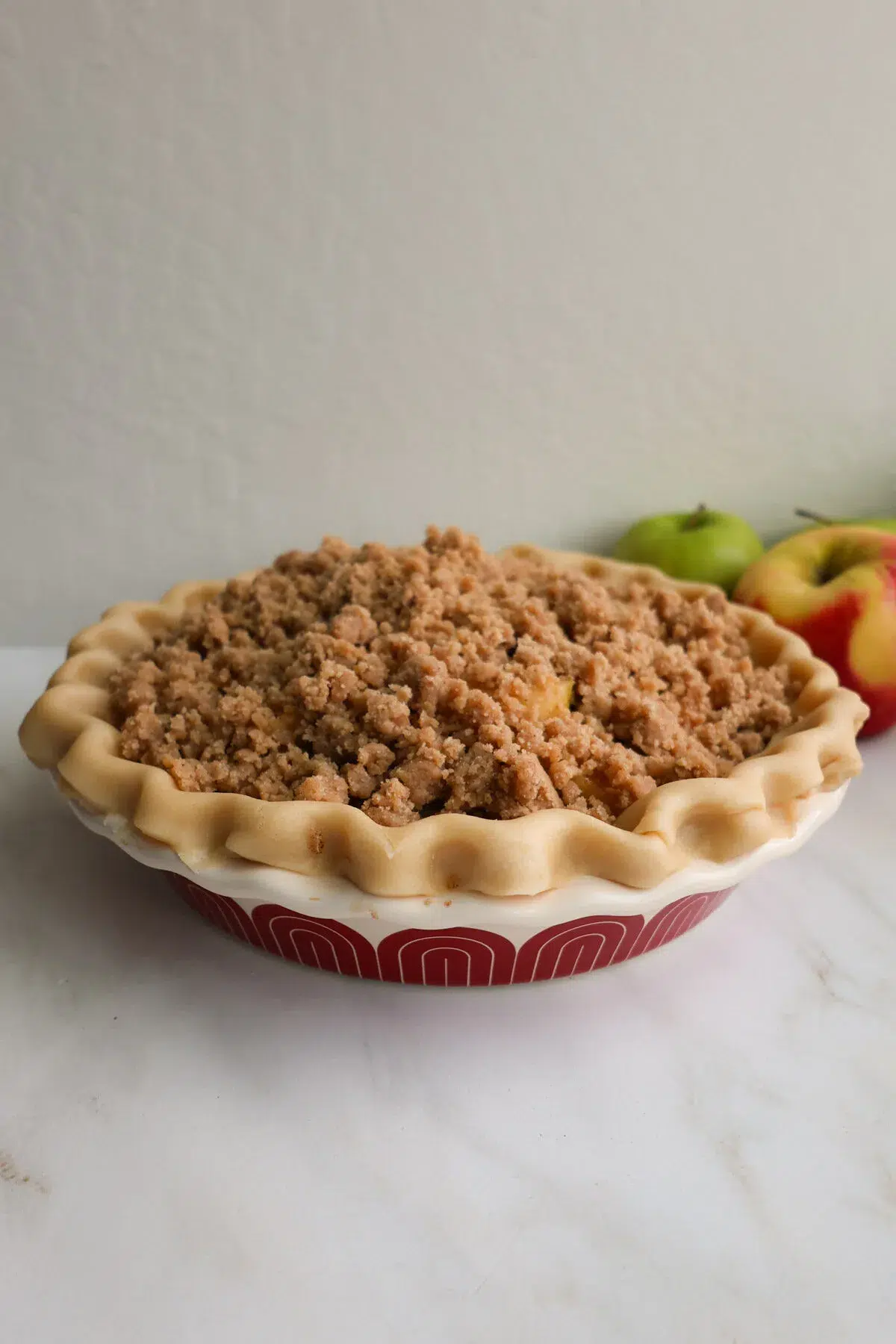 An unbaked apple crumble pie, topped with crumb topping, and two apples next to it.