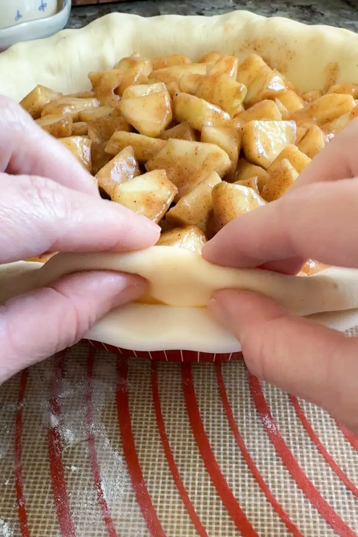 Two hands folding the edge of the crust of an unbaked apple pie crust underneath.