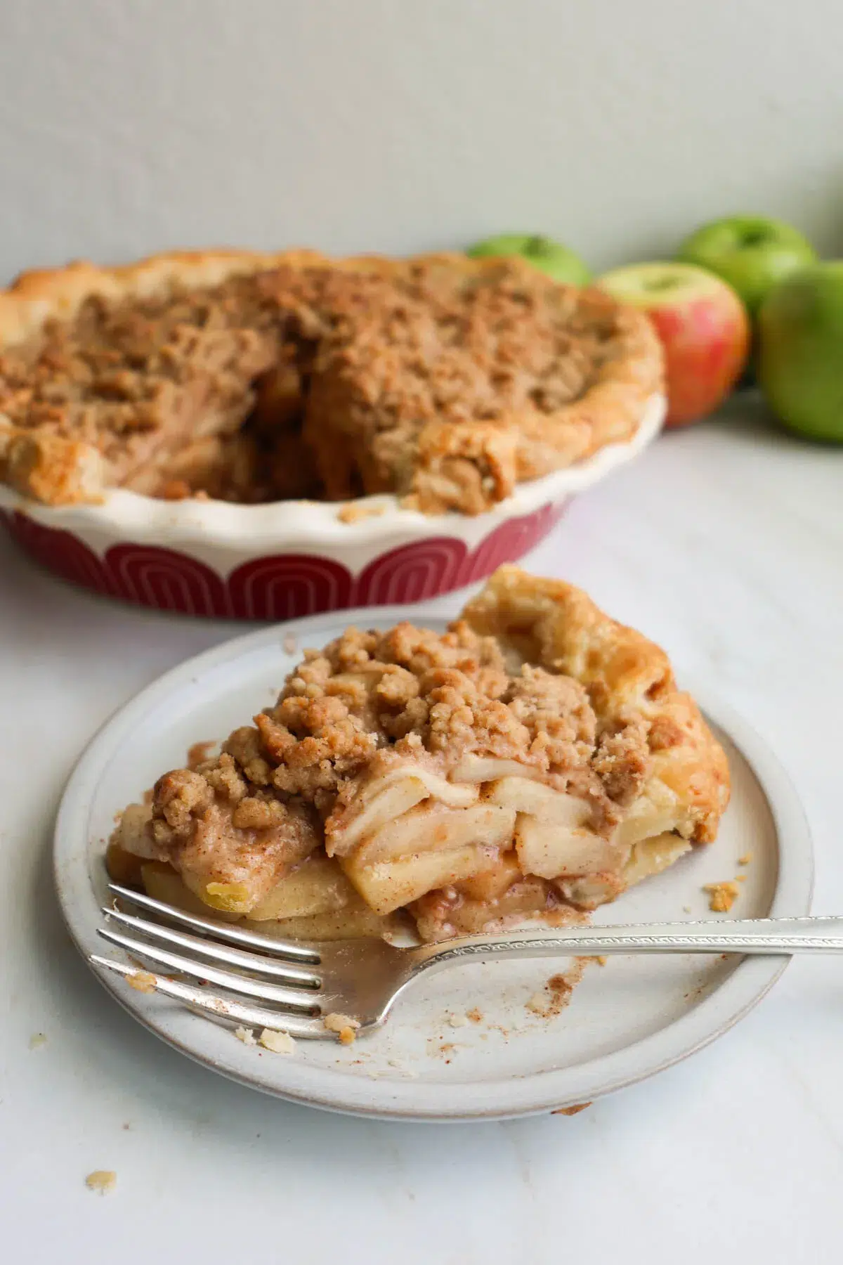 A slice of apple crumble pie, on a dessert plate with a fork, and a pie behind it with a slice taken out.