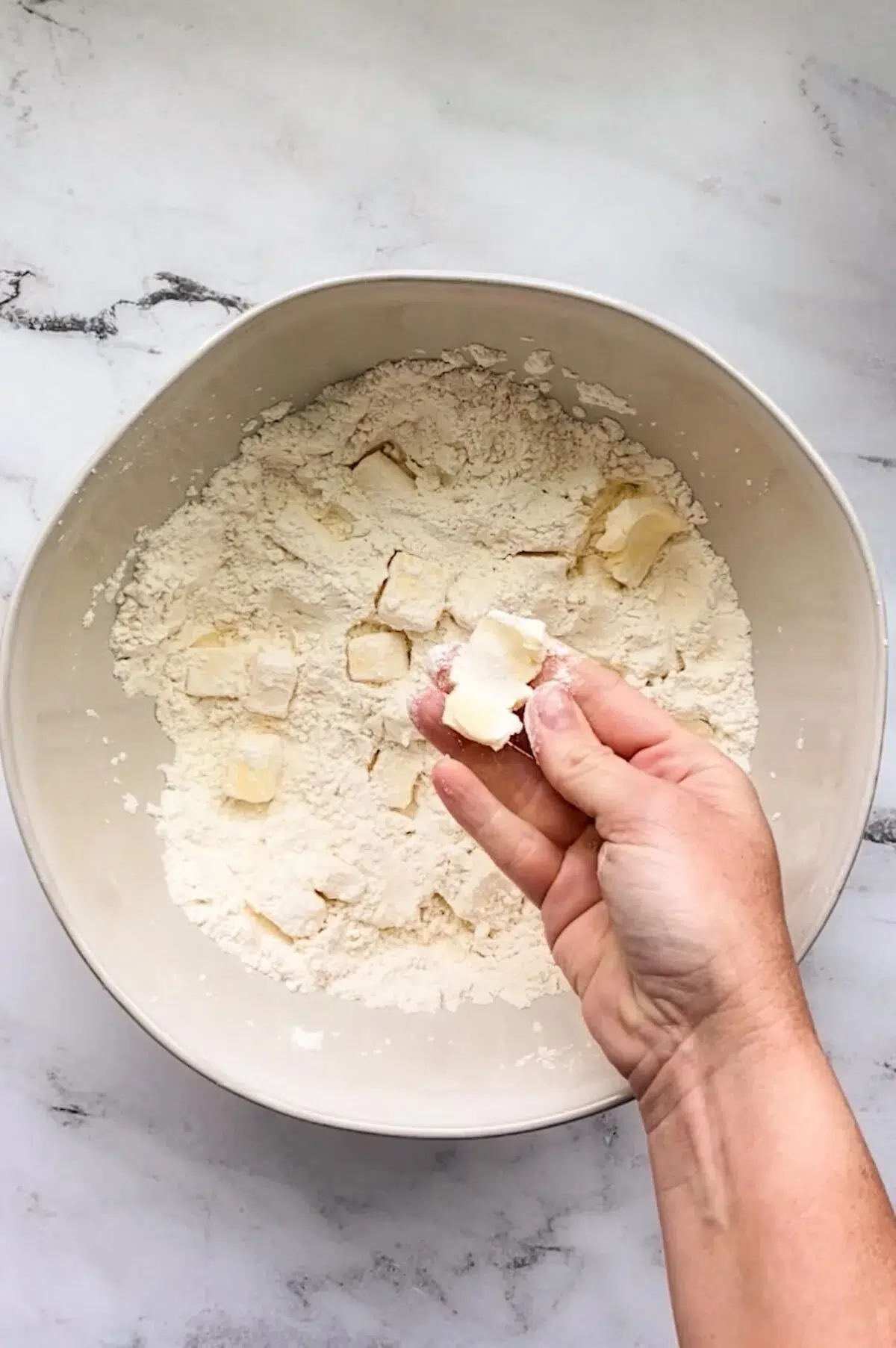 A bowl with the ingredients for butter pie crust, and a hand holding a flattened cube of butter.