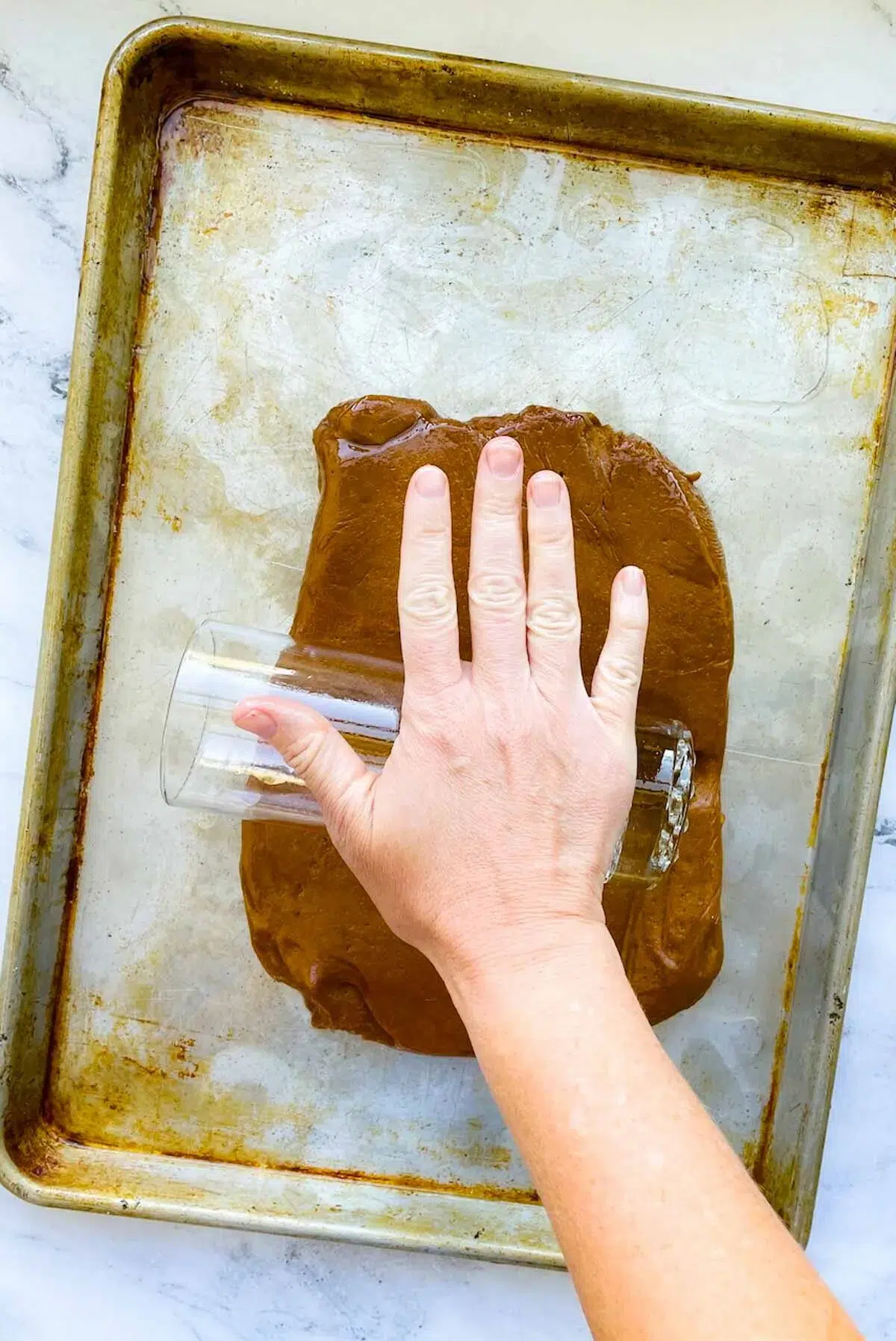 A hand rolling a tall glass cup over a disc of gingerbread dough, on an oiled baking sheet.