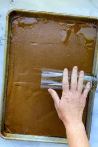 Gingerbread house dough, rolled out to fill a half sheet baking sheet, with a hand rolling a long glass cup over the dough.