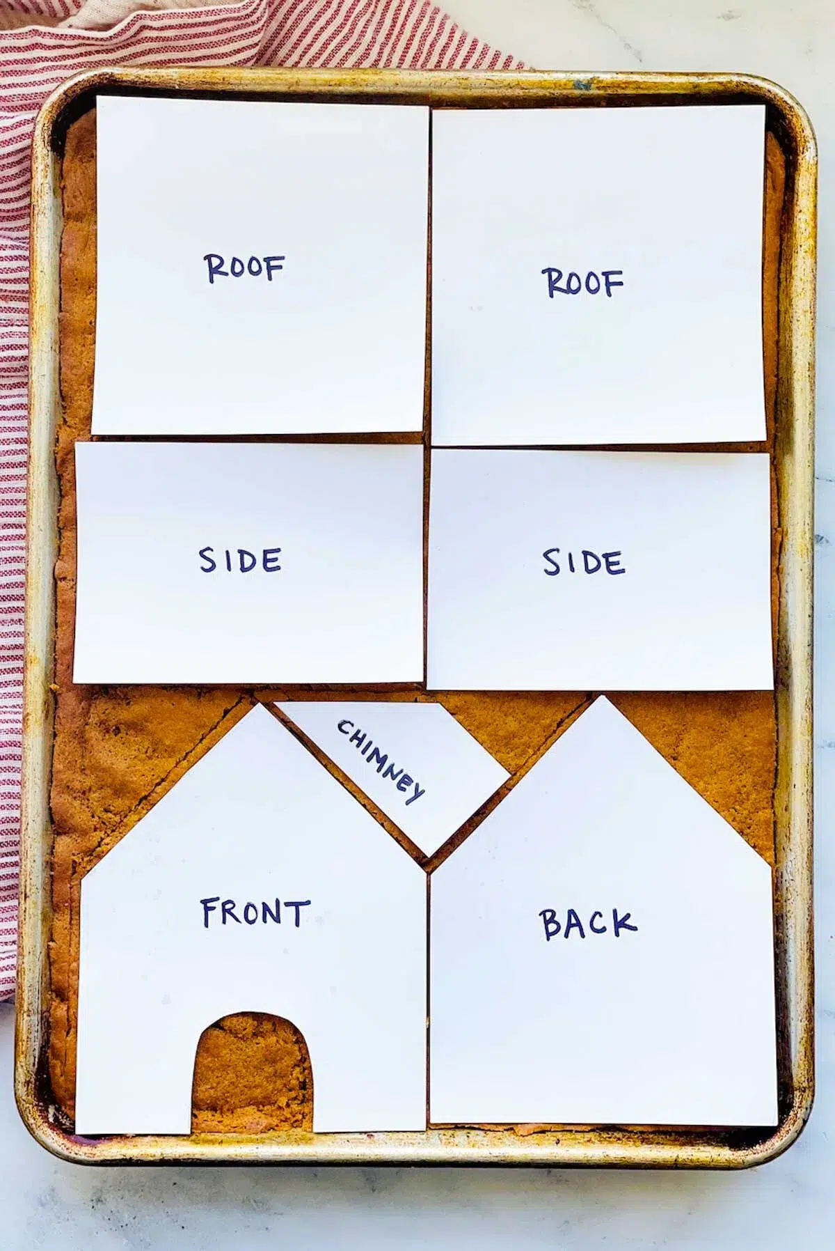 The gingerbread house template cutouts arranged over the baked gingerbread, with the roof pieces at the top, then the side pieces, and then the front and back pieces, with the chimney piece in the empty space above the front piece.