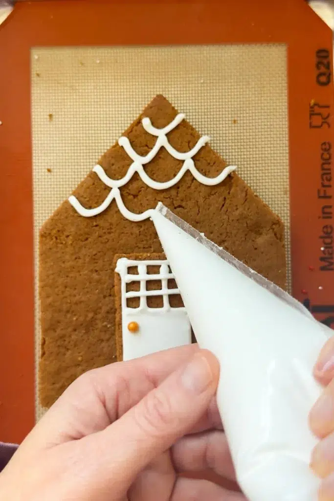 A front gingerbread cutout laying on a baking sheet lined with a silpat mat, and two hands holding a piping bag full of royal icing, piping designs onto the gingerbread.