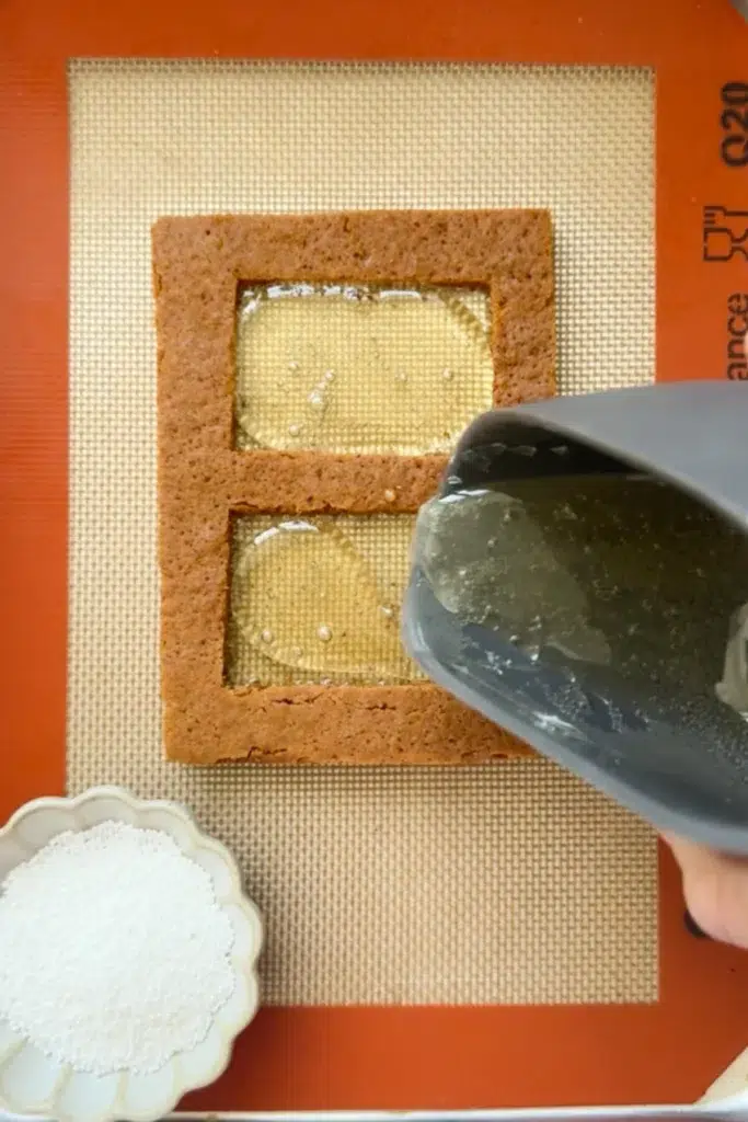 A gingerbread house cut out, resting on a silicone baking mat, with two windows cut out of it and a bowl of melted isomalt being poured into the empty window spaces.
