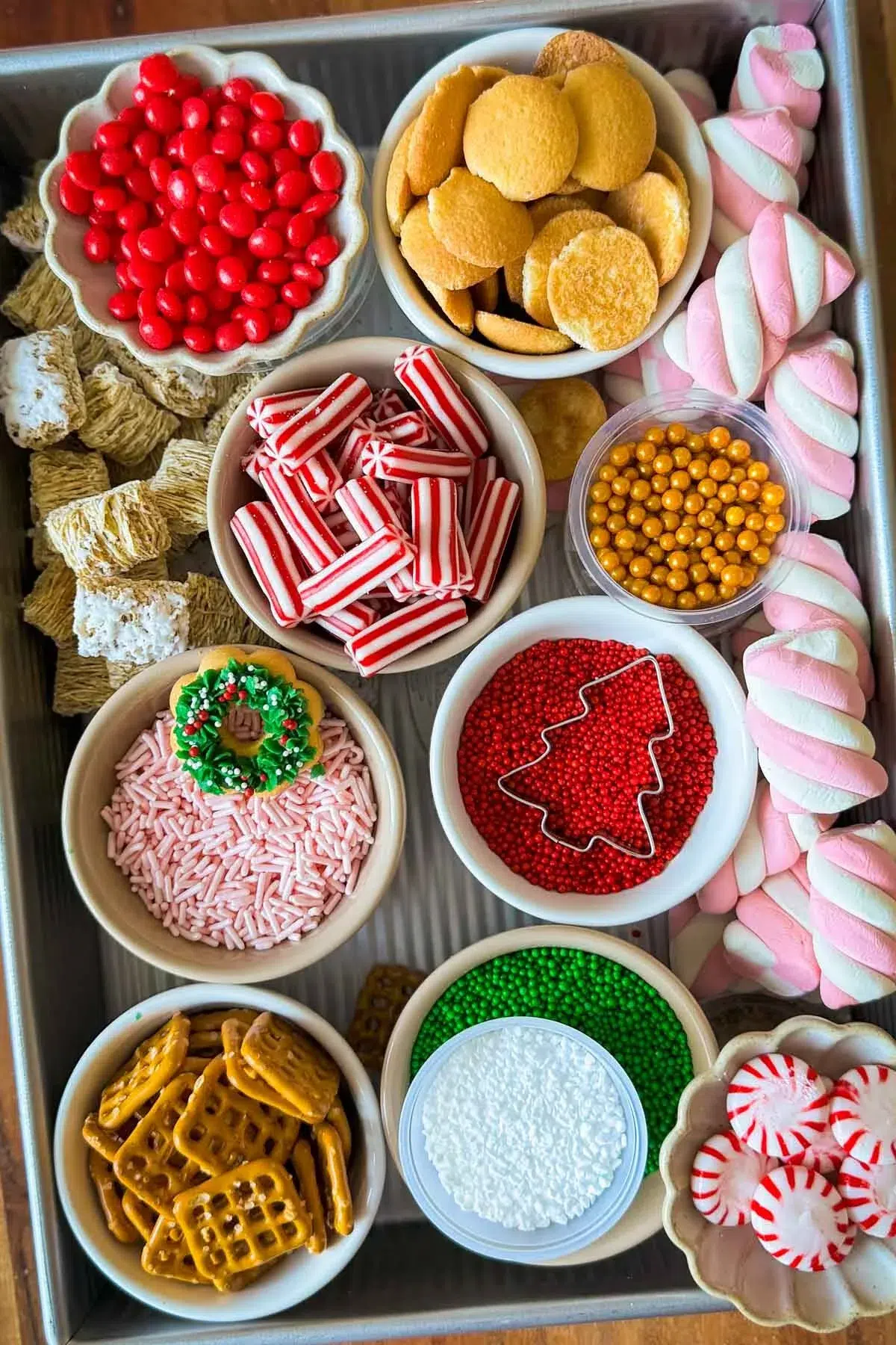 A cake pan full of bowls containing gingerbread house decorations including sprinkles, peppermints, pretzels, Nilla wafers, and marshmallows.