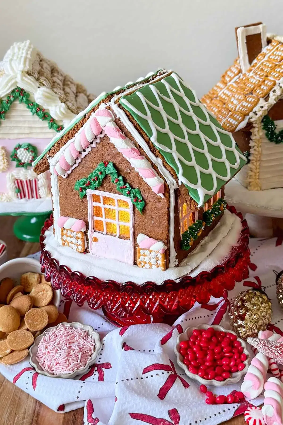 A decorated gingerbread house on a cake platter, with sugar around the base of the house, two more decorated gingerbread house behind it, and candy and cookies on the table beside it.