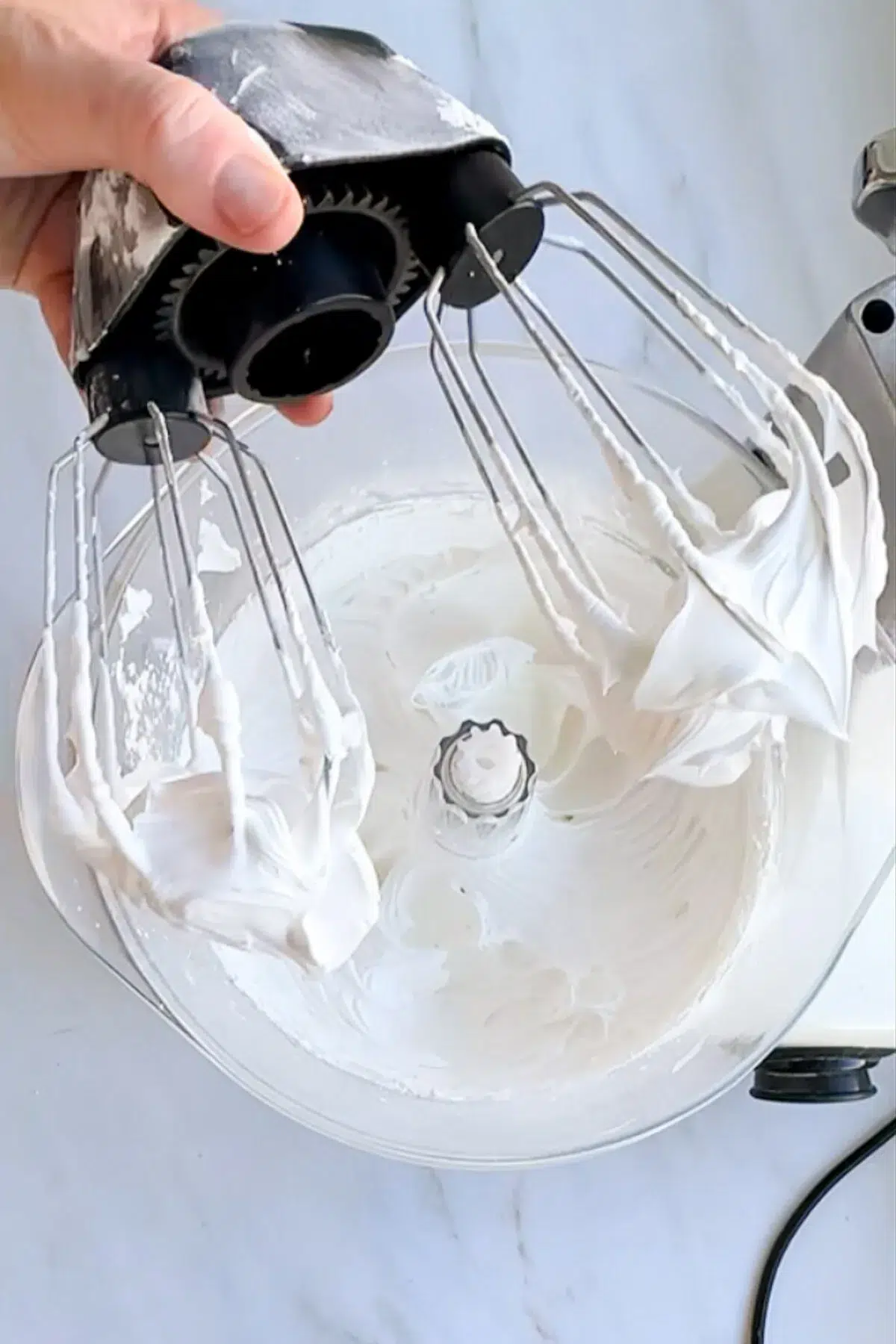 A hand holding a wire whisk attachment, with stiff royal icing on the whisks, and the mixer bowl underneath it with more royal icing.