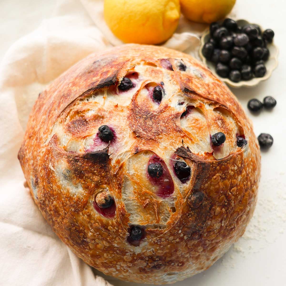 A baked loaf of lemon sourdough bread, on a linen napkin, with lemons and blueberries surrounding it.