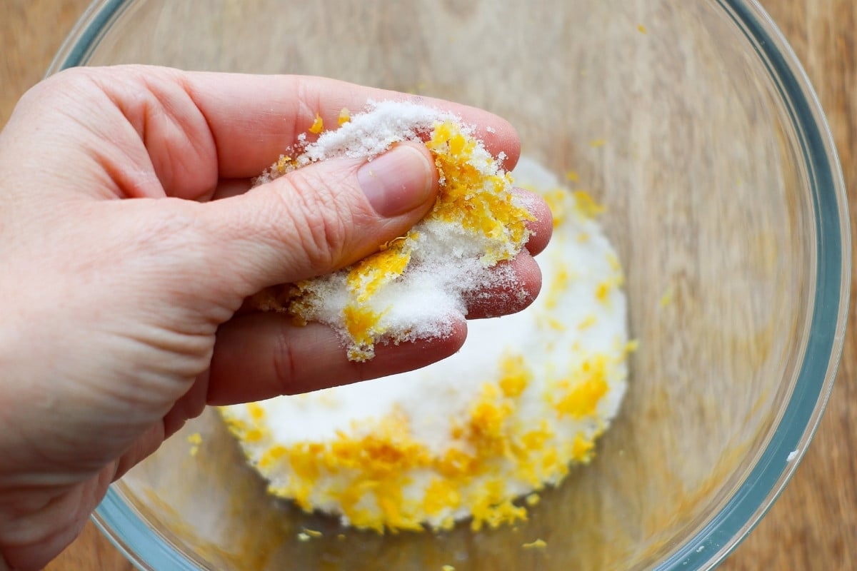 A hand rubbing sugar and lemon zest together over a glass bowl containing more sugar and lemon zest.