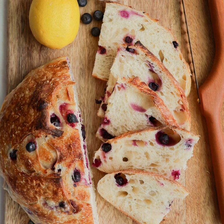 A loaf of lemon blueberry sourdough bread, with half of it sliced, on a a wood cutting board.