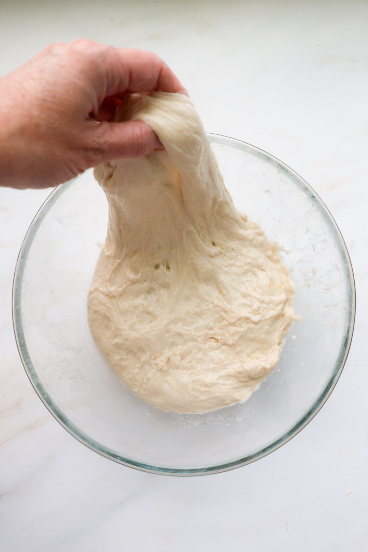 A hand stretching sourdough dough up as it rests in a glass bowl.