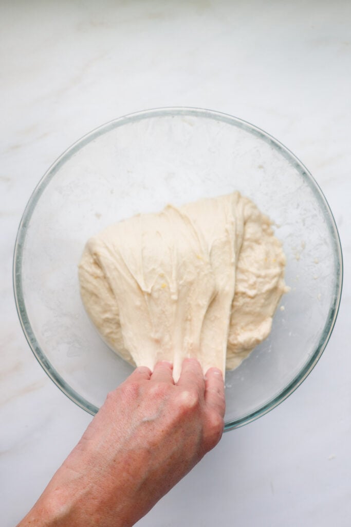 A hand folding sourdough over onto itself while it's resting in a glass bowl.