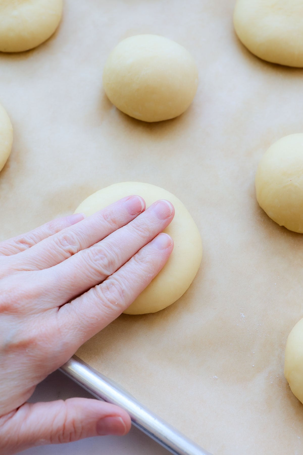 A hand flattening a piece of brioche dough on a baking sheet lined with parchment, and other dough balls on it.