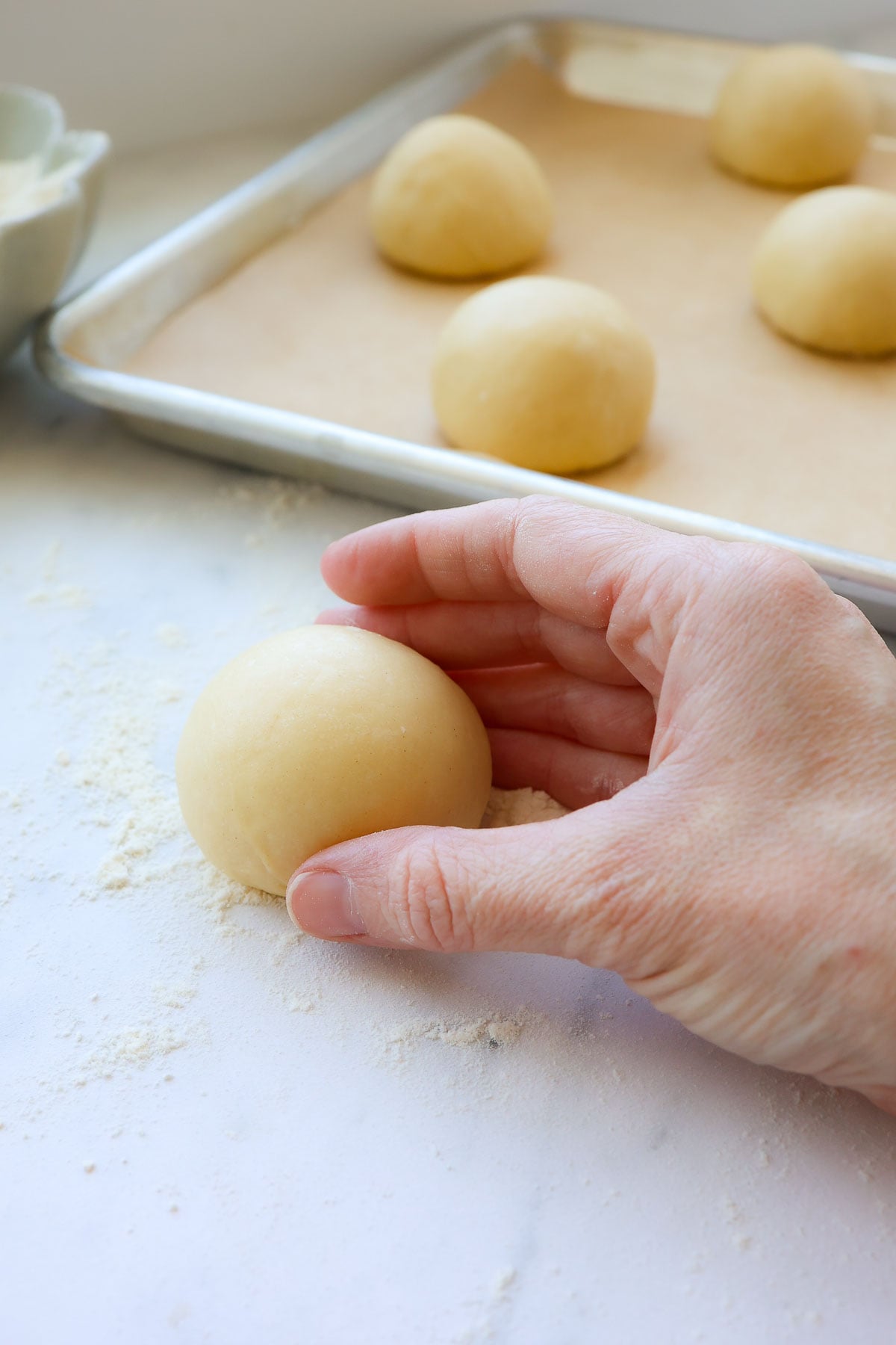 A hand rolling a brioche dough ball and more dough balls on a baking sheet with parchment paper.