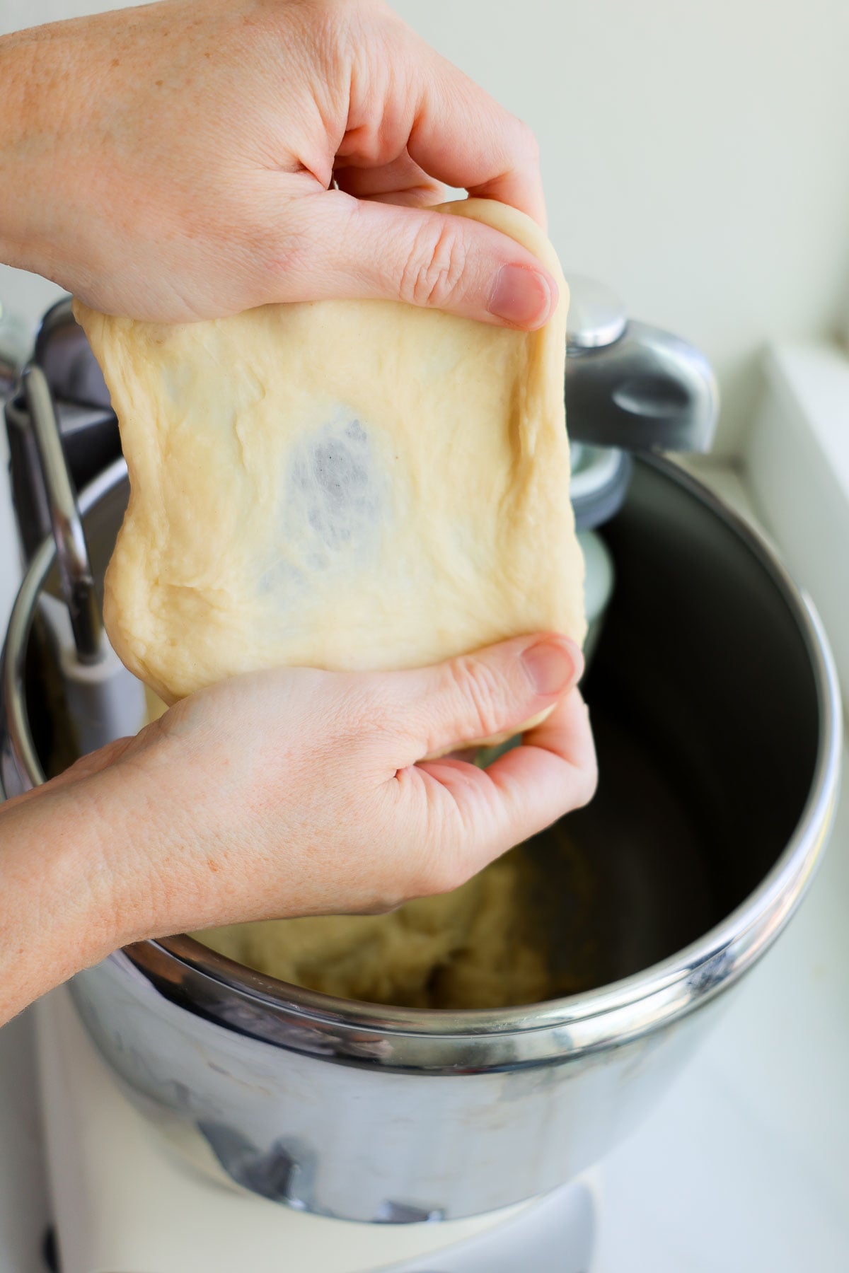 Two hands pulling apart a piece of brioche bun dough, with the center translucent like a windowpane.