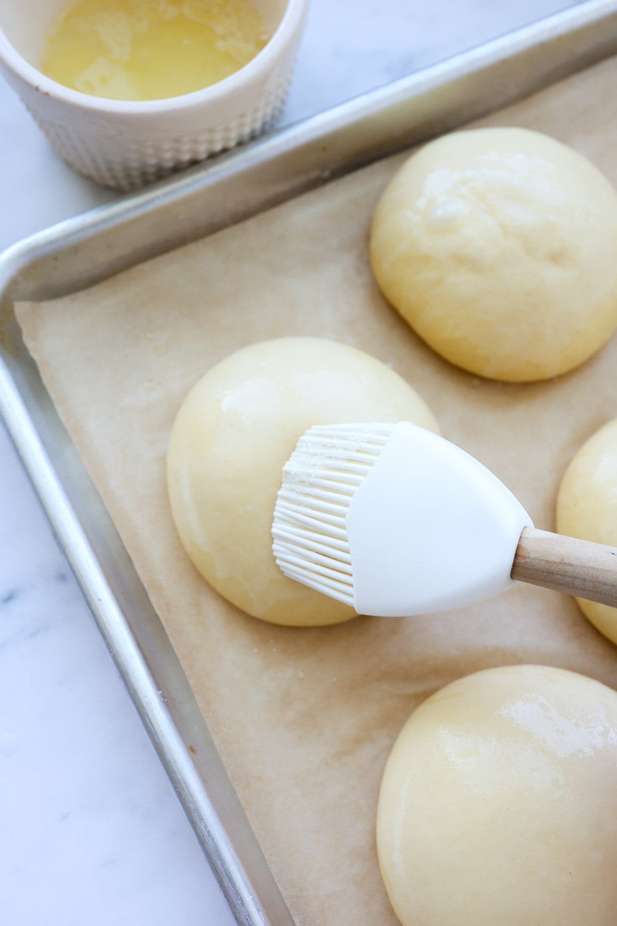 A ball of brioche hamburger bun dough, being brushed with egg wash, with a silicone pastry brush.